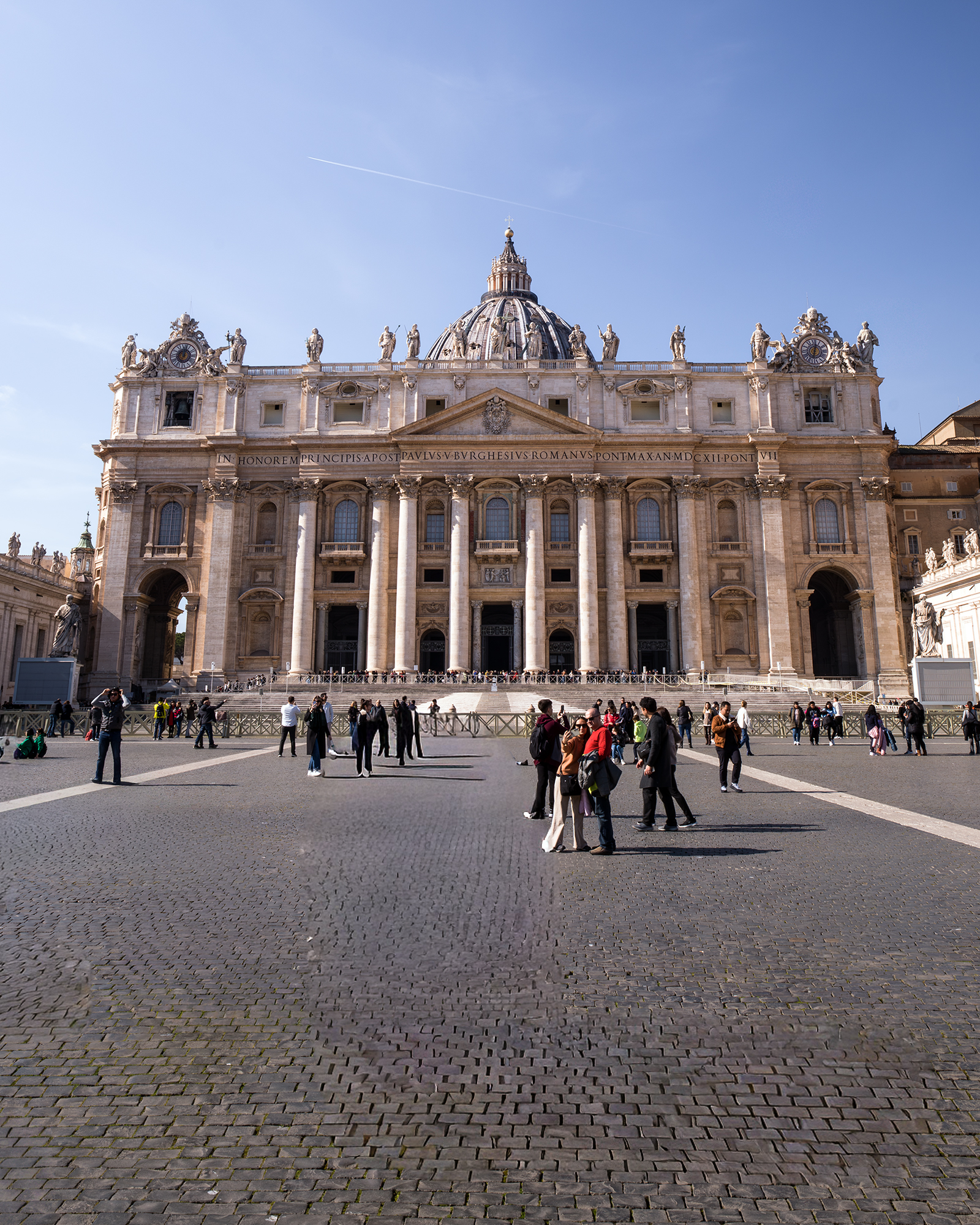 Vatican - An ornate basilica facade with a central dome rises above tourists scattered across a wide cobblestone plaza in bright sunlight. - #architecture #basilica #dome #plaza #travel #tourists #statues #cobblestone #sunlight #historic