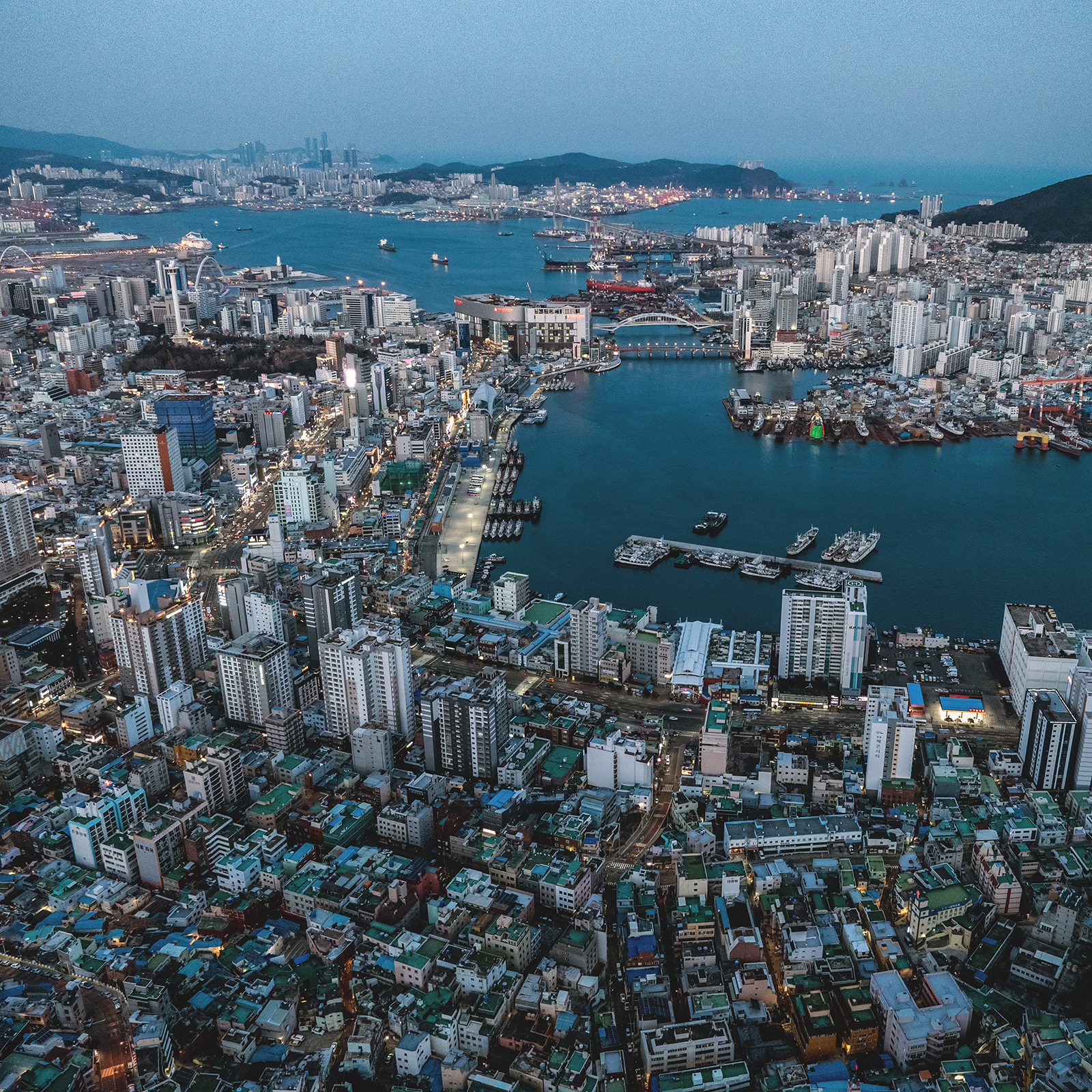 Busan - Aerial view of a dense coastal city around a harbor, with high-rises, bridges, boats, and glowing streets under cool blue evening light. - #Busan #BlueHour #BusanHarbor #Cityscape #UrbanPhotography #TravelPhotography #NightPhotography #KoreaTravel #VisitBusan #HarborView