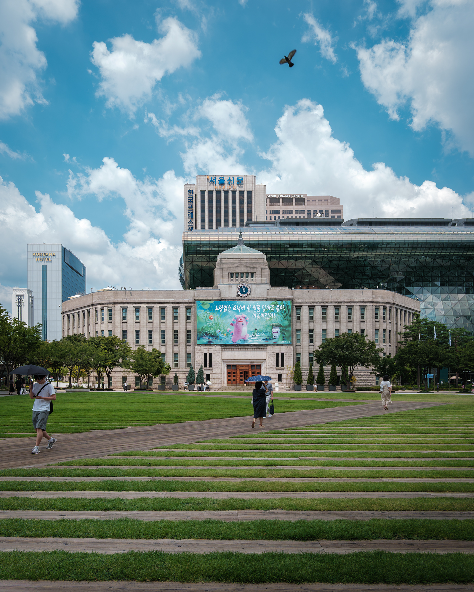 Seoul City Hall - A civic building with a clock and large banner faces a green lawn, with pedestrians under umbrellas beneath bright clouds and a blue sky. - #SeoulCityHall #Seoul #VisitSeoul #SouthKorea #Korea #SeoulArchitecture #ModernArchitecture #TravelPhotography #Cityscape #UrbanPhotography