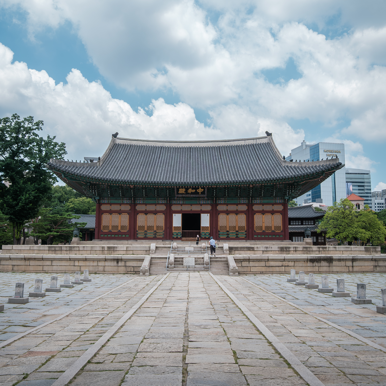 Deoksugung Palace - A traditional palace hall stands beyond a broad stone courtyard under bright clouds, with modern buildings and trees framing the background. - #Deoksugung #DeoksugungPalace #Seoul #VisitSeoul #Korea #SouthKorea #JoseonDynasty #KoreanPalace #RoyalPalace #HistoricSite