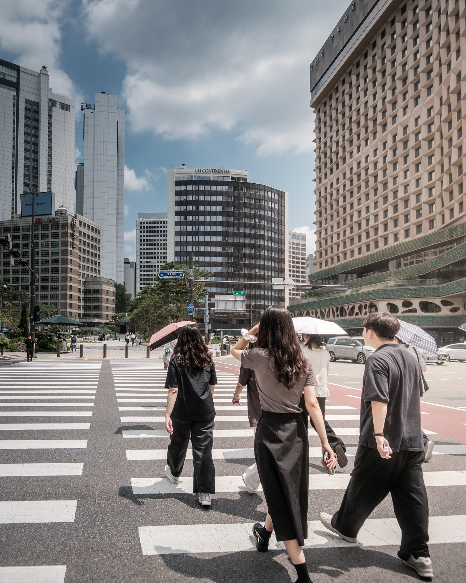 Crosswalk - Pedestrians cross a wide city crosswalk under bright midday light, framed by tall buildings and a partly cloudy sky. - #Seoul #Crosswalk #SeoulCrosswalk #PedestrianCrossing #StreetPhotography #UrbanPhotography #Cityscape #CityLife #SeoulStreets #UrbanLife