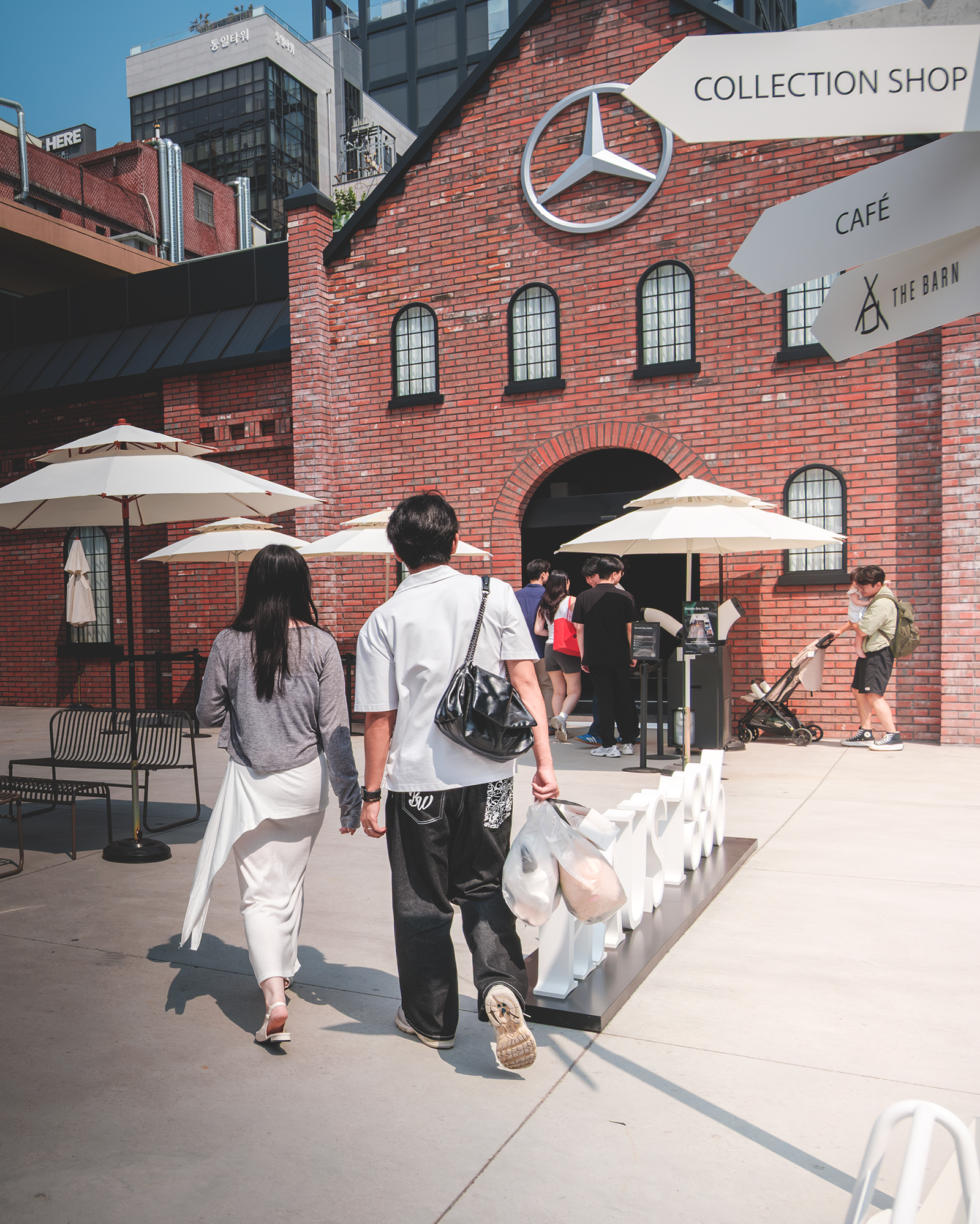Couple - Two people walk toward a sunlit red-brick storefront with white umbrellas and city buildings in the background. - #StreetPhotography #UrbanScene #BrickBuilding #SunnyDay #CityLife #Architecture #Pedestrians #OutdoorCafe #RedBrick #Candid