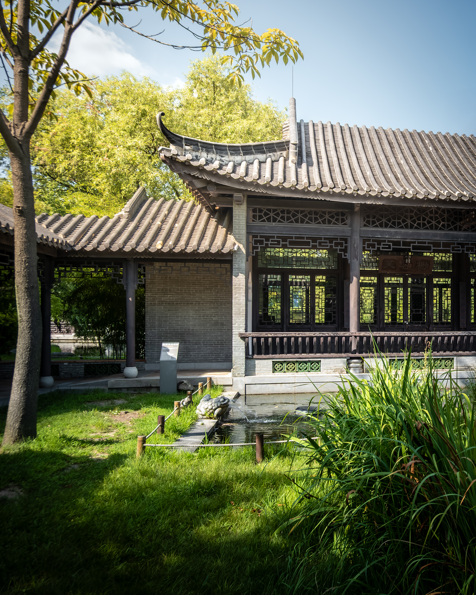 Wolhwawon - A traditional garden pavilion with tiled roofs and lattice windows stands beside a small pond amid lush greenery in bright sunlight. - #Wolhwawon #WolhwawonGarden #Suwon #SuwonKorea #HyowonPark #GyeonggiDo #SouthKorea #KoreaTravel #VisitKorea #TravelKorea