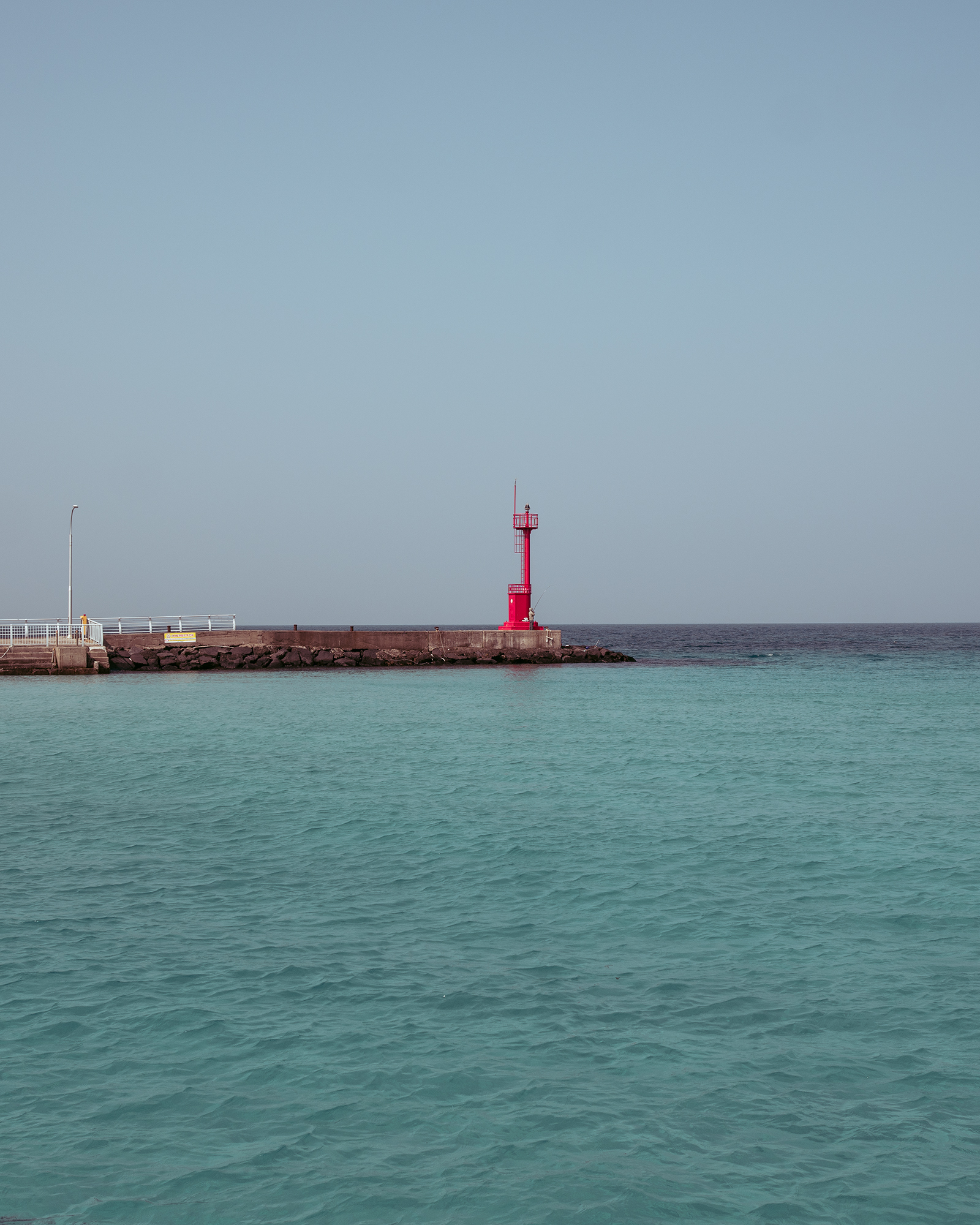 Gimnyeong Sea - A red beacon stands on a rocky breakwater beside turquoise water under a wide, pale blue sky. - #Gimnyeong #GimnyeongBeach #GimnyeongSea #Jeju #JejuIsland #JejuBeach #JejuSea #Korea #SouthKorea #KoreanBeach