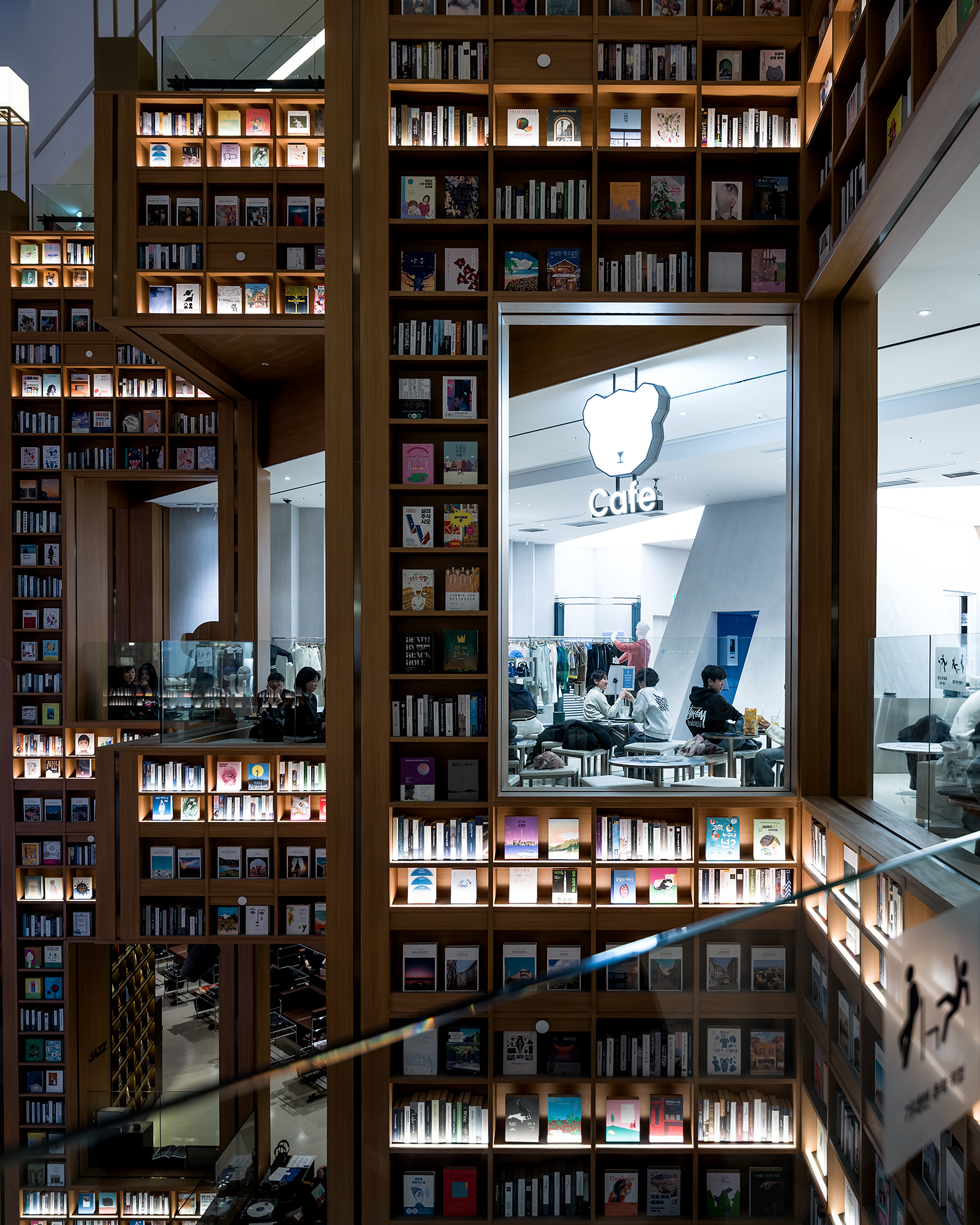Starfield Library - Tall wooden bookshelves glow with warm lights around a bright cafe window, creating layered geometric views inside a modern library. - #StarfieldLibrary #StarfieldSuwon #Suwon #SuwonKorea #SuwonTravel #Gyeonggi #GyeonggiDo #Korea #KoreaTravel #VisitKorea