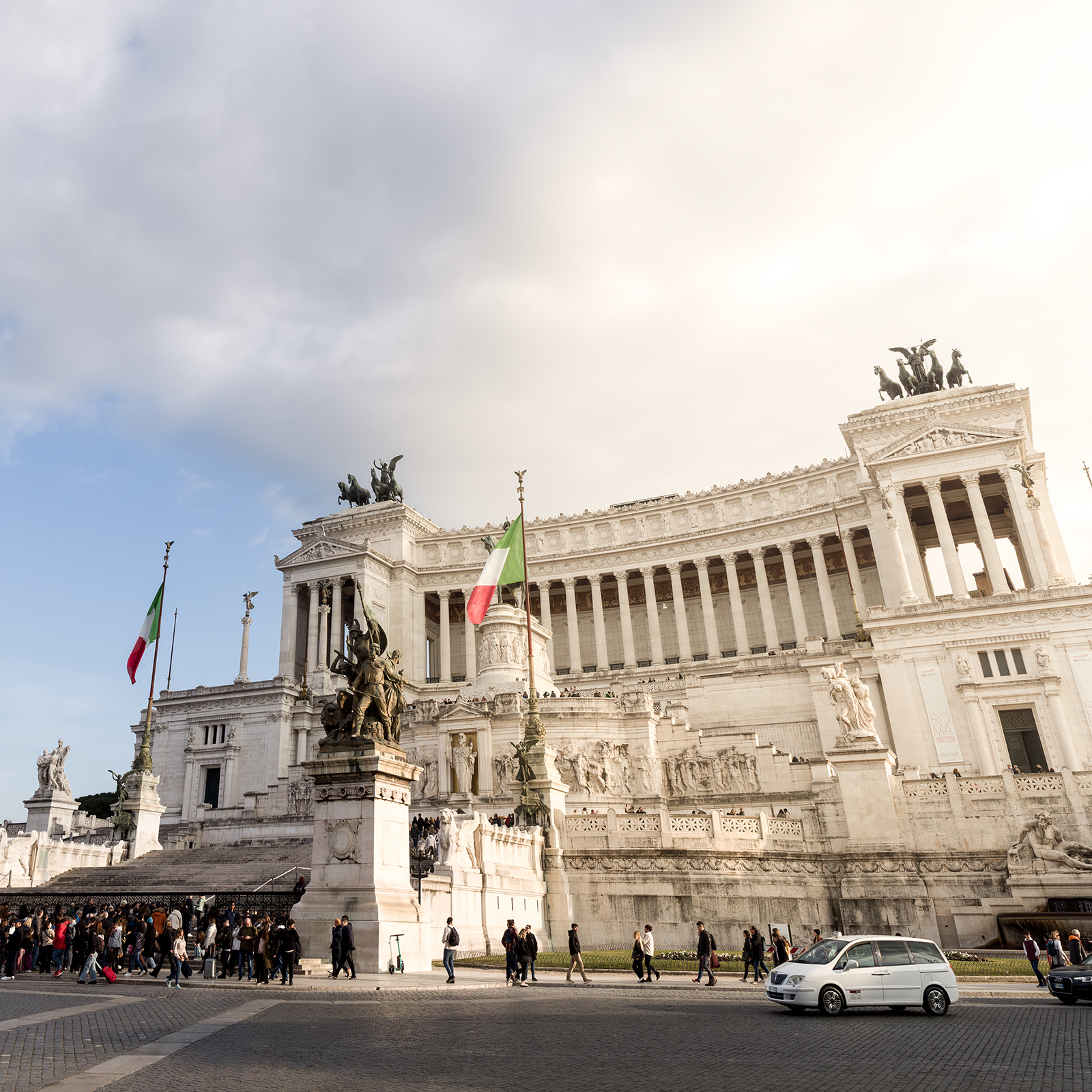 Rome - A sunlit white monument with columns, statues, Italian flags, pedestrians, and cars stands beneath a wide cloudy sky. - #Rome #Roma #Italy #RomeItaly #VisitRome #RomeTravel #TravelRome #ExploreRome #ItalyTravel #VisitItaly