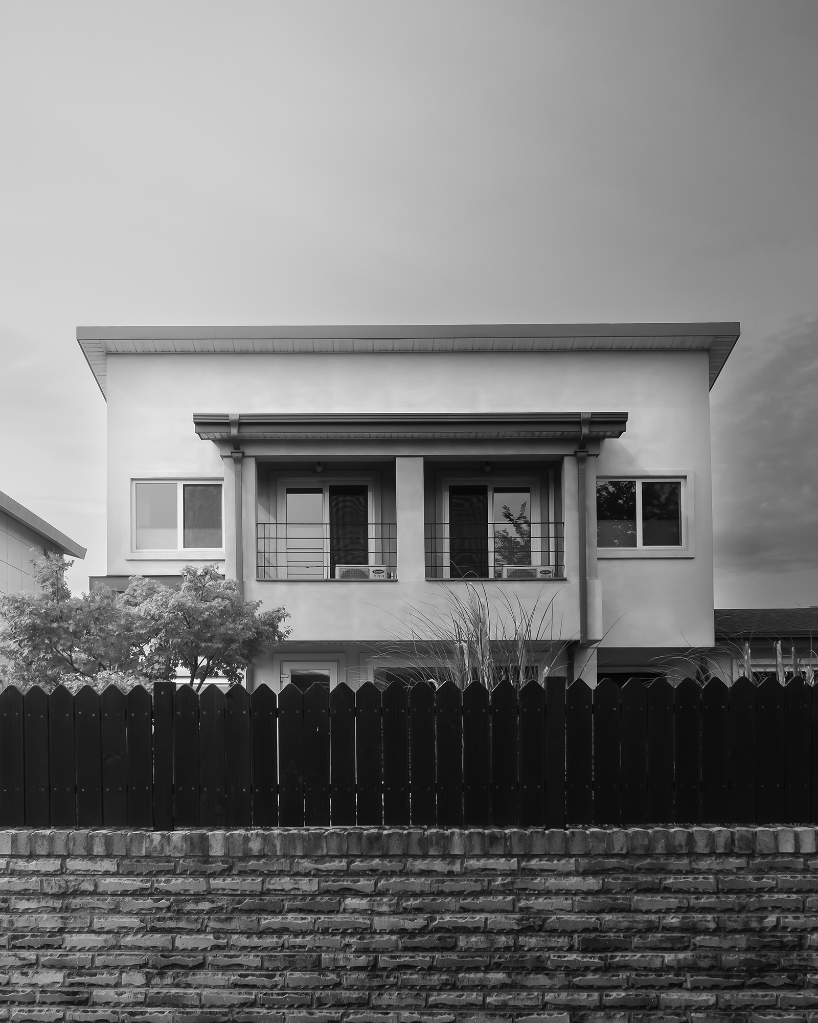 House - A black-and-white view shows a modern two-story house behind a dark picket fence and stone wall under a cloudy sky. - #Home #House #PrivateHouse #ResidentialHouse #Residence #ResidentialArea #Neighborhood #HomeExterior #HousePhotography #Architecture