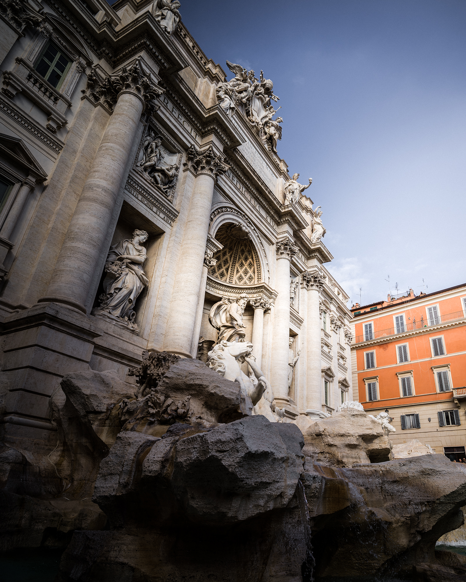 Rome - A low-angle view shows an ornate stone fountain facade with statues, columns, rocky water features, and a warm orange building under a clear blue sky. - #Rome #Roma #Italy #RomeItaly #VisitRome #RomeTravel #TravelRome #ExploreRome #ItalyTravel #VisitItaly