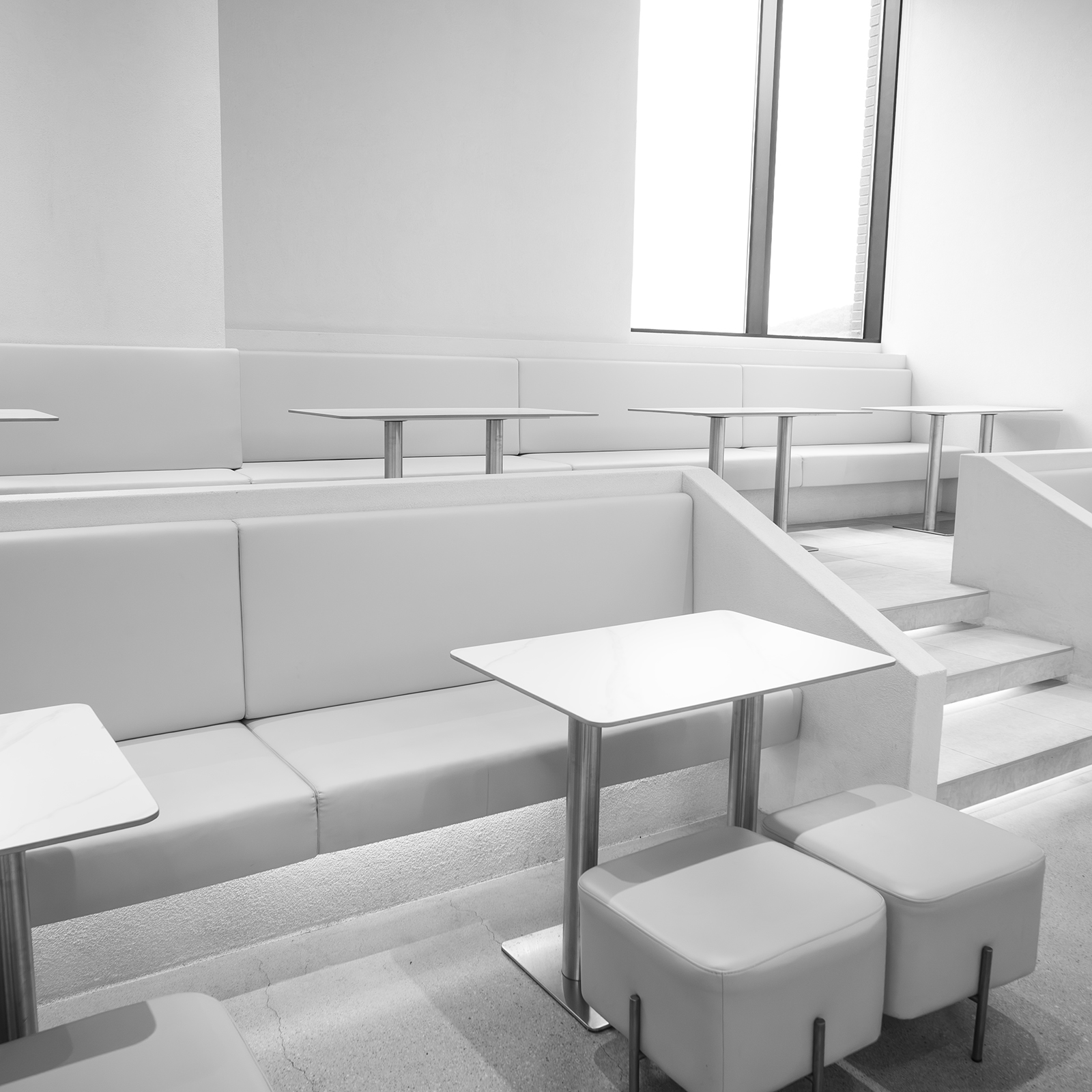 Table - Bright monochrome seating area with white tables, padded benches, stools, steps, and a tall window in a minimalist interior. - #CafeTable #Cafe #CoffeeShop #CoffeeTime #CafeVibes #CafeLife #CafeInterior #CoffeeLover #CoffeeCulture #CozyCafe