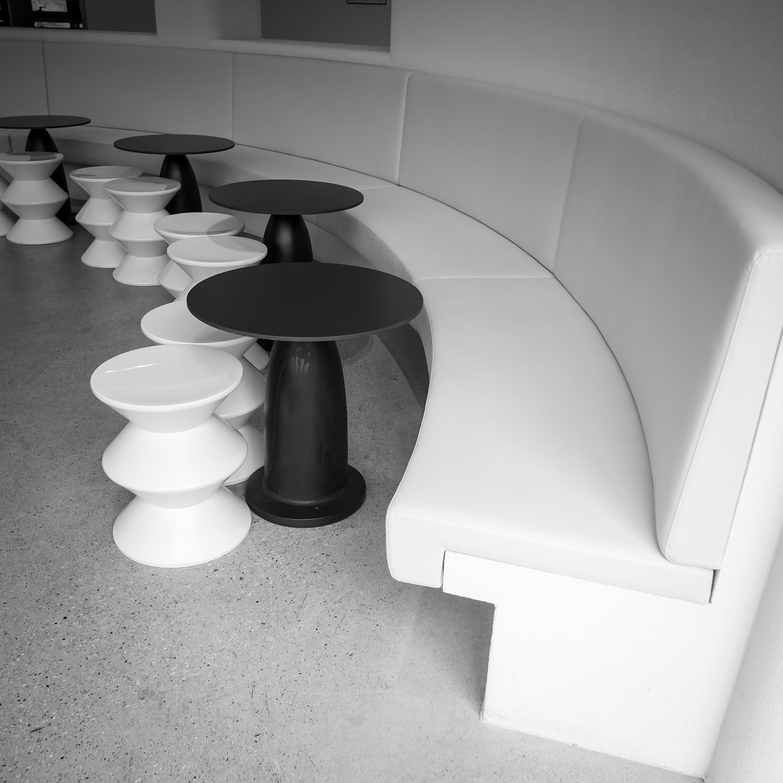 Table - A black-and-white view shows curved white bench seating with round black tables and white stools in a minimalist interior. - #CafeTable #Cafe #CoffeeShop #CoffeeTime #CafeVibes #CafeLife #CafeInterior #CoffeeLover #CoffeeCulture #CozyCafe