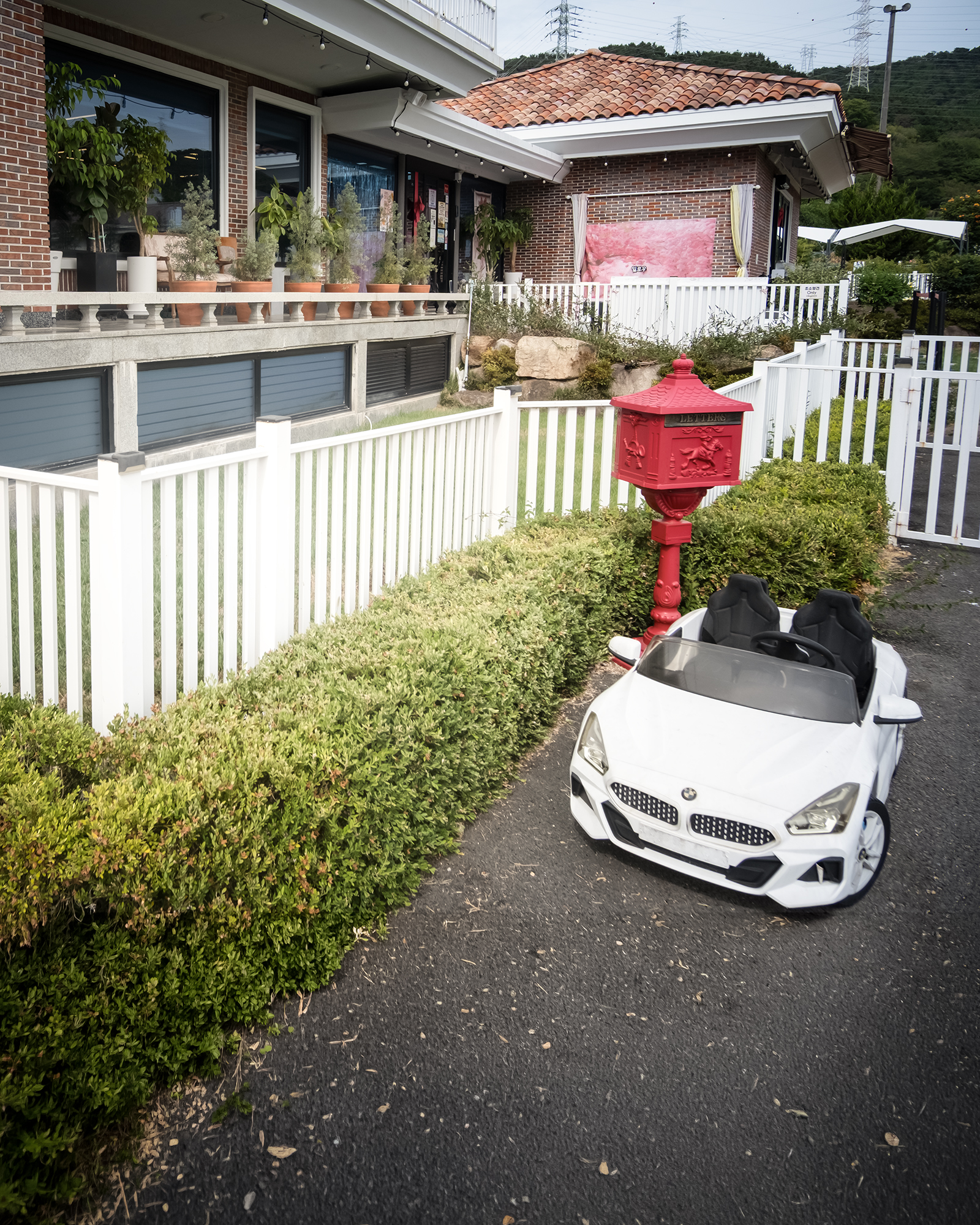 Kids' Car - A white children's ride-on car sits beside a red mailbox, trimmed hedges, and a white fence outside a brick building. - #KidsCar #ChildrensCar #RideOnCar #KidsRideOnCar #ToyCar #KidsVehicle #ChildrensToy #KidsToy #Playtime #KidsPlay