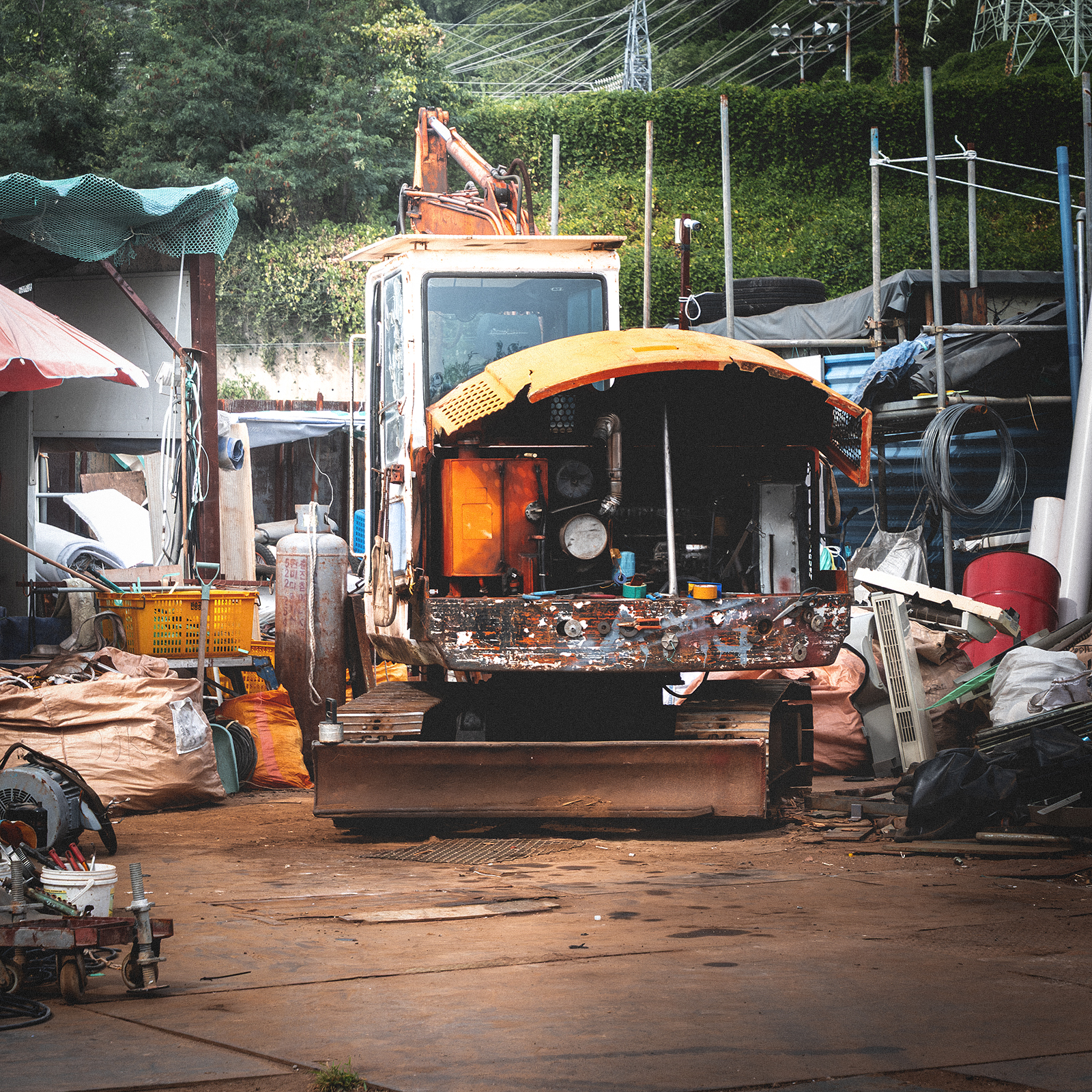 Junkyard - A weathered orange excavator sits with its front panel open amid scattered scrap and tools in a cluttered yard under soft daylight. - #Junkyard #ScrapYard #Scrapyard #JunkShop #ScrapMetal #MetalScrap #OldThings #Abandoned #Industrial #IndustrialPhotography