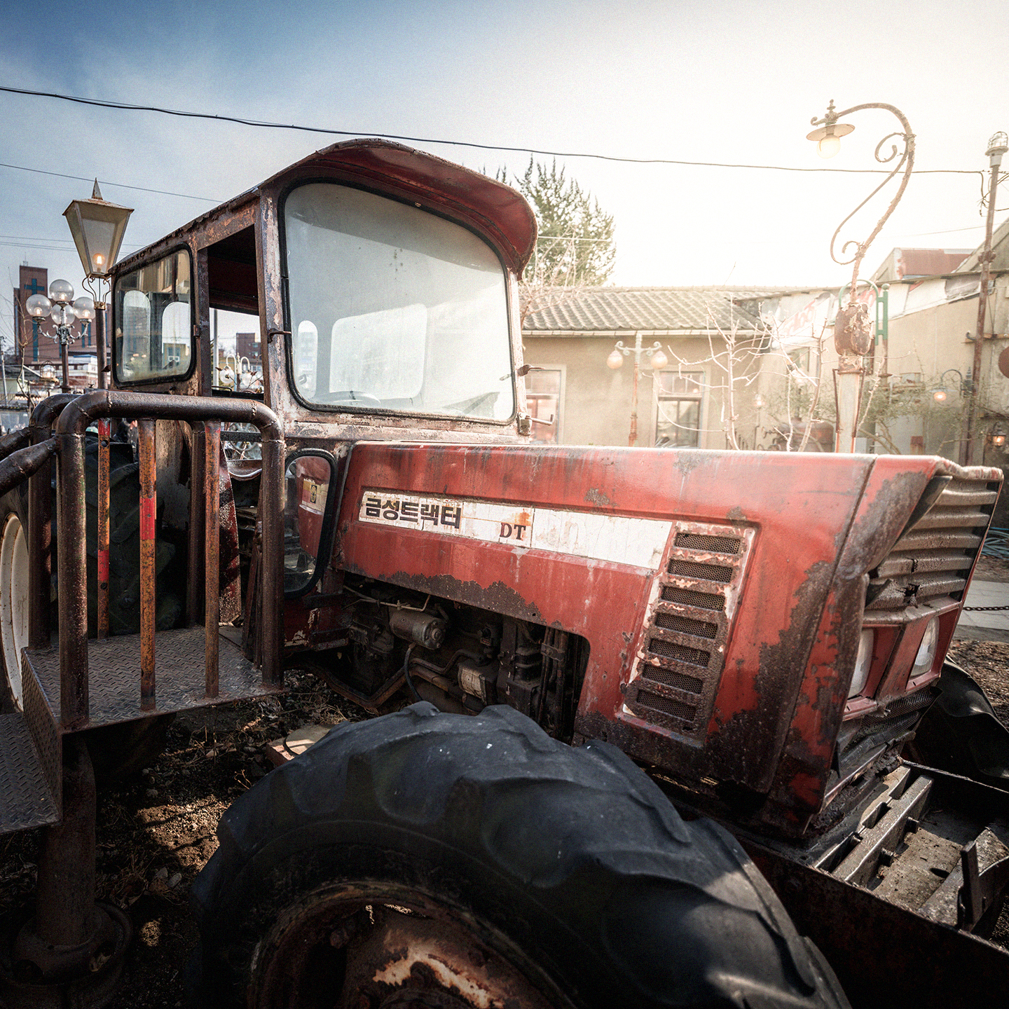 Tractor - A weathered red tractor fills the frame in bright sunlight, with vintage lamps and low buildings behind it. - #Tractor #FarmTractor #AgriculturalTractor #Farming #Agriculture #FarmLife #FarmMachinery #FarmEquipment #TractorLife #TractorPhotography