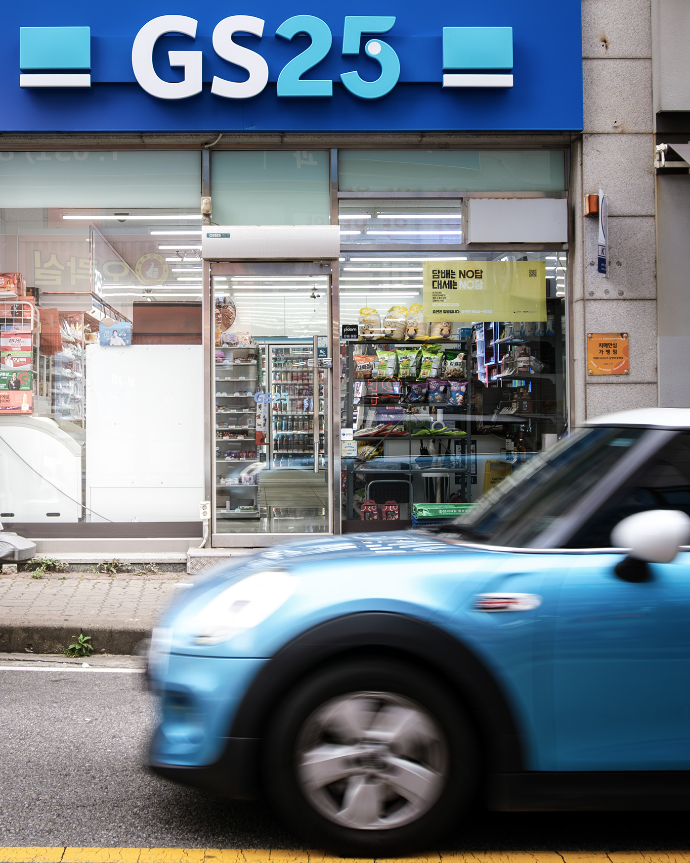 GS25 - A blue car blurs past a brightly lit GS25 storefront, framing the shop entrance with urban motion and glass reflections. - #ConvenienceStore #KoreanConvenienceStore #ConvenienceShop #CornerStore #Korea #KoreanLife #DailyLife #EverydayKorea #StreetPhotography #UrbanPhotography