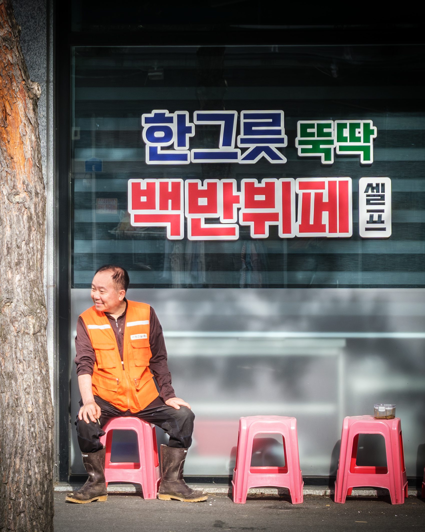 Worker - A man in an orange safety vest sits on a pink stool beneath bold Korean window lettering in bright street light. - #Worker #Workers #Laborer #Labor #WorkingPeople #WorkLife #AtWork #HardWorking #Workplace #DailyWork