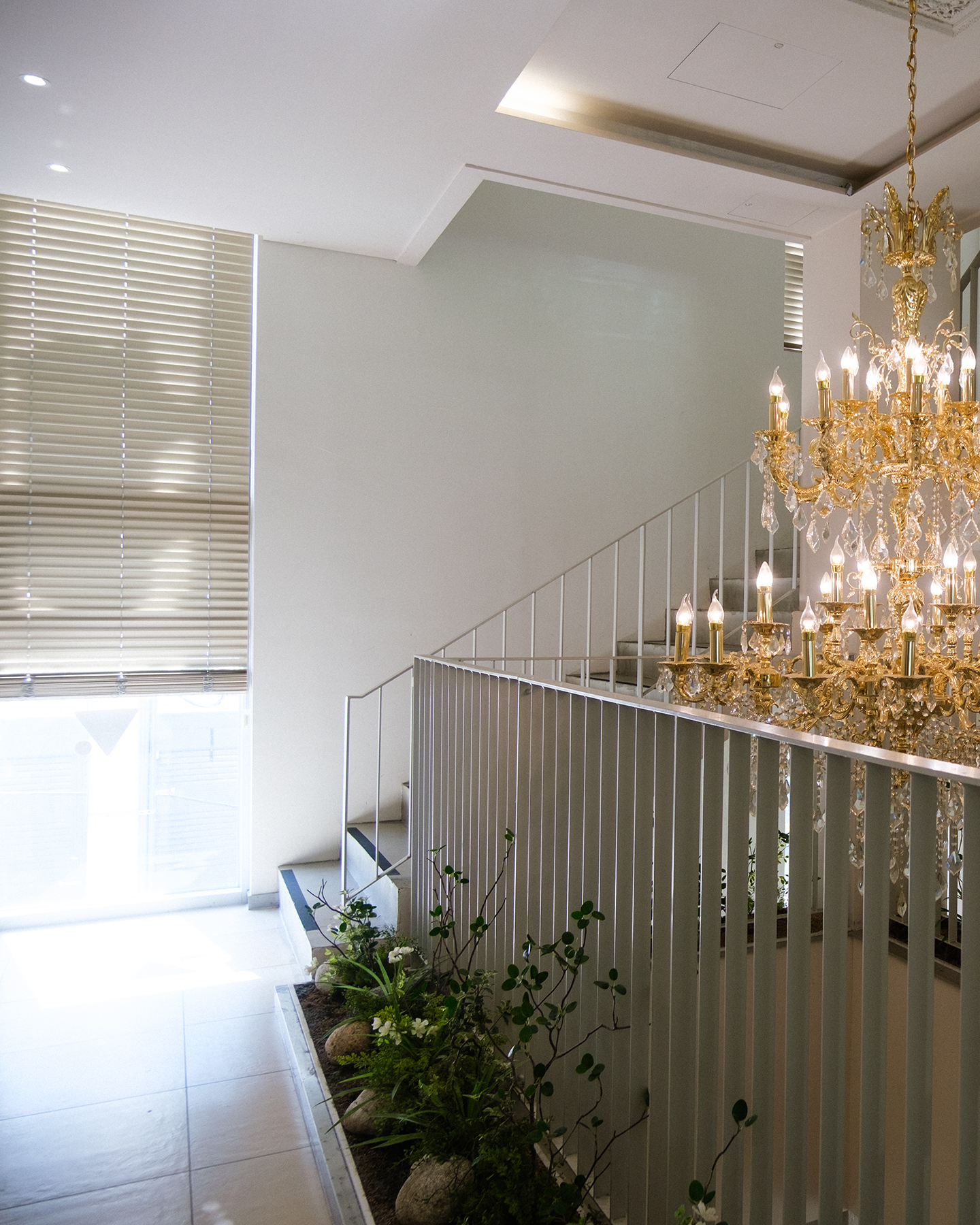 Chandelier - A glowing crystal chandelier hangs beside a staircase, with window blinds diffusing soft daylight across the bright interior. - #Cafe #Chandelier #Table #HouseYongam #SouthKorea