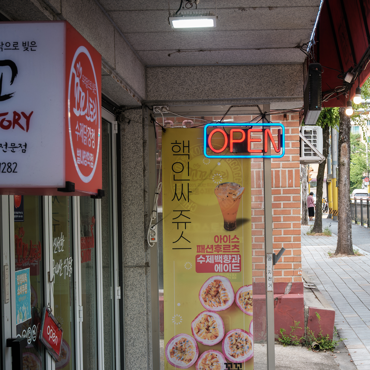 Open - A neon OPEN sign hangs above a fruit juice poster at a small storefront beside a quiet sidewalk. - #Store #Street #Juice