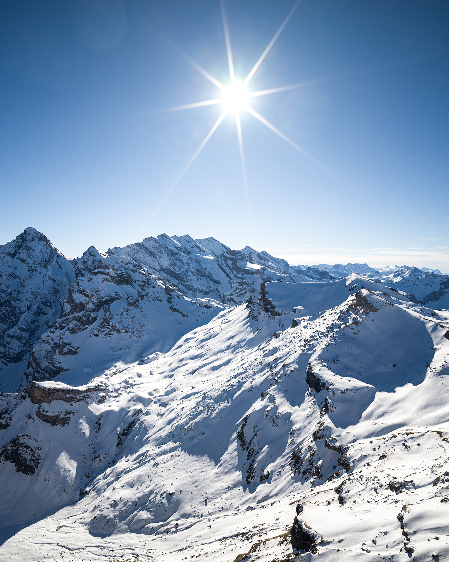 Schilthorn - A bright sunburst shines over snow-covered alpine peaks beneath a clear blue sky, casting crisp shadows across the rugged slopes. - #Schilthorn #Switzerland #SwissAlps #BerneseOberland #Murren #AlpineViews #MountainViews #SwissMountains #Alps #TravelSwitzerland