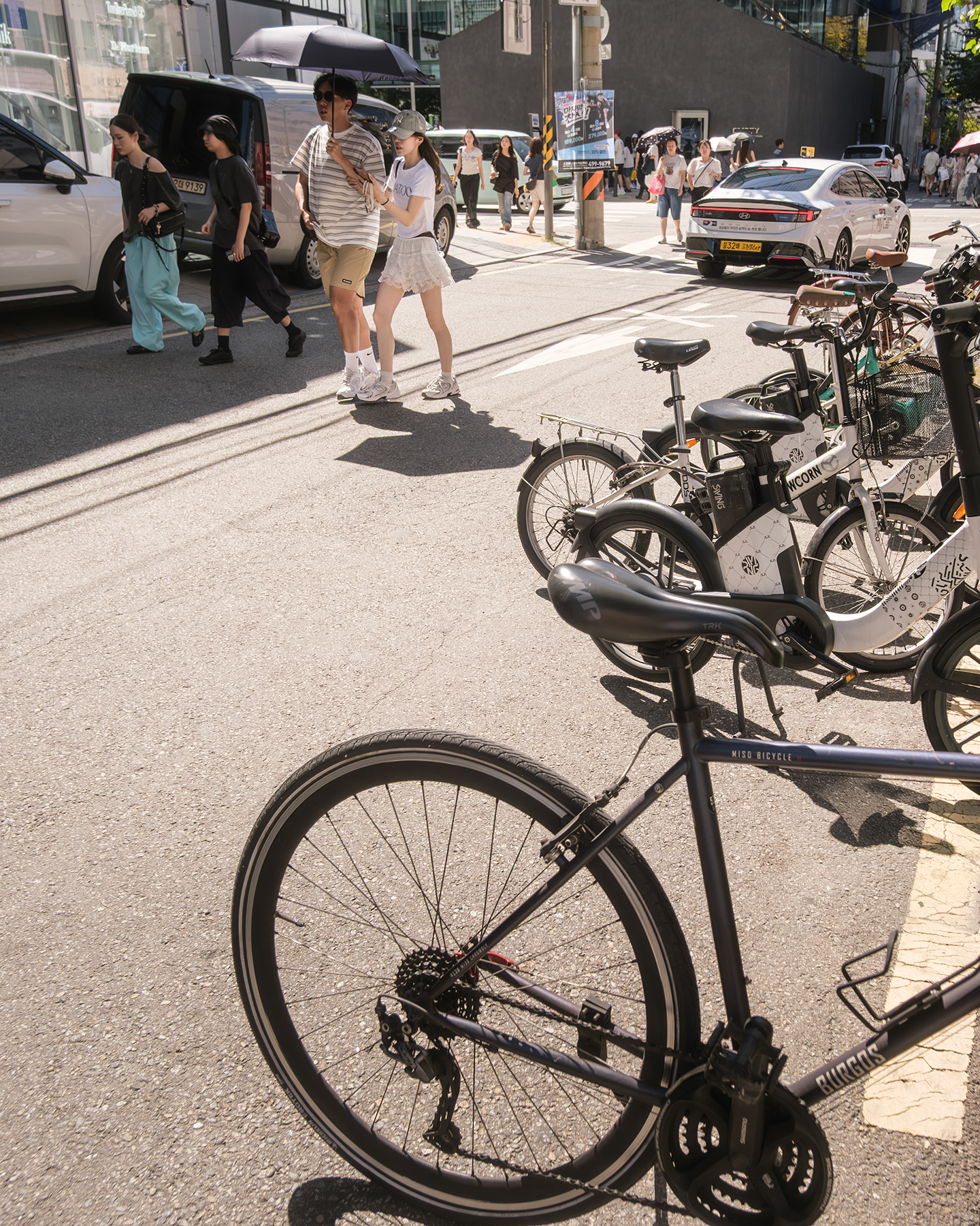 Bicycle - Parked bicycles fill the foreground as pedestrians with umbrellas cross a sunlit city street. - #Bicycle #Bike #Street #Pedestrian #Passerby #StreetLife #StreetScene #StreetPhotography #UrbanLife #UrbanStreet