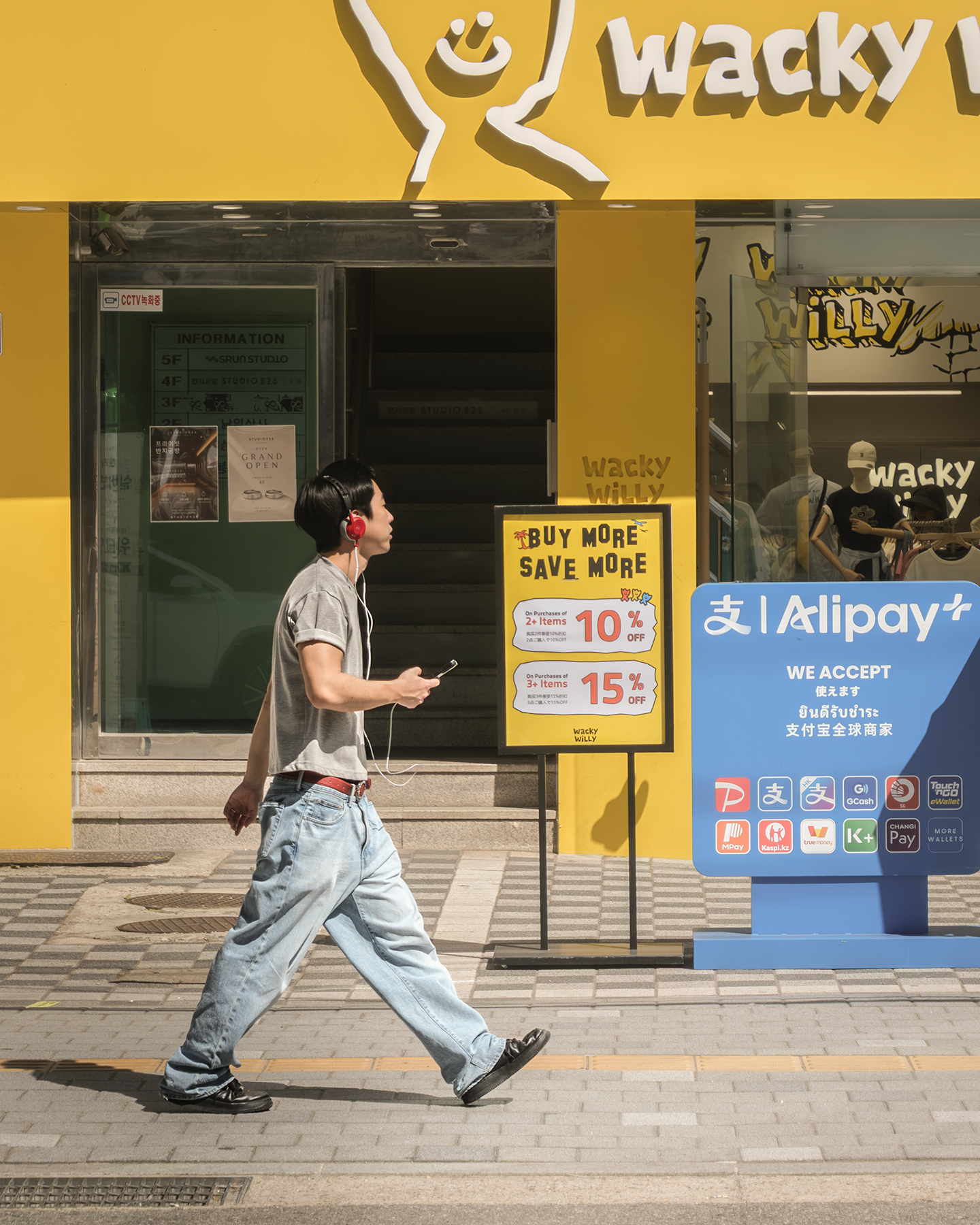 Music - A pedestrian with red headphones walks past a bright yellow storefront and sidewalk signs in strong sunlight. - #Music #SeongsuDong #Street #Seoul #SouthKorea