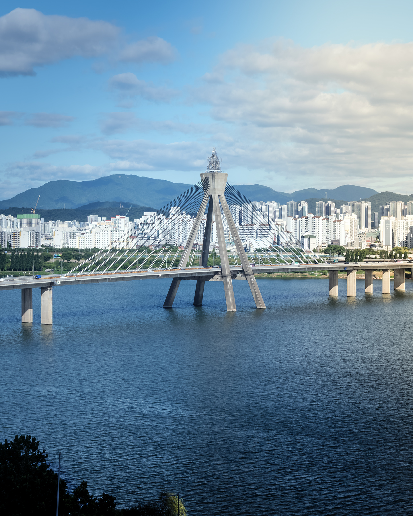 Han River - A cable-stayed bridge spans a broad river before high-rise buildings, green mountains, and a partly cloudy blue sky. - #HanRiver #Hangang #Seoul #SouthKorea #Korea #SeoulCity #SeoulLife #SeoulTravel #VisitSeoul #ExploreSeoul