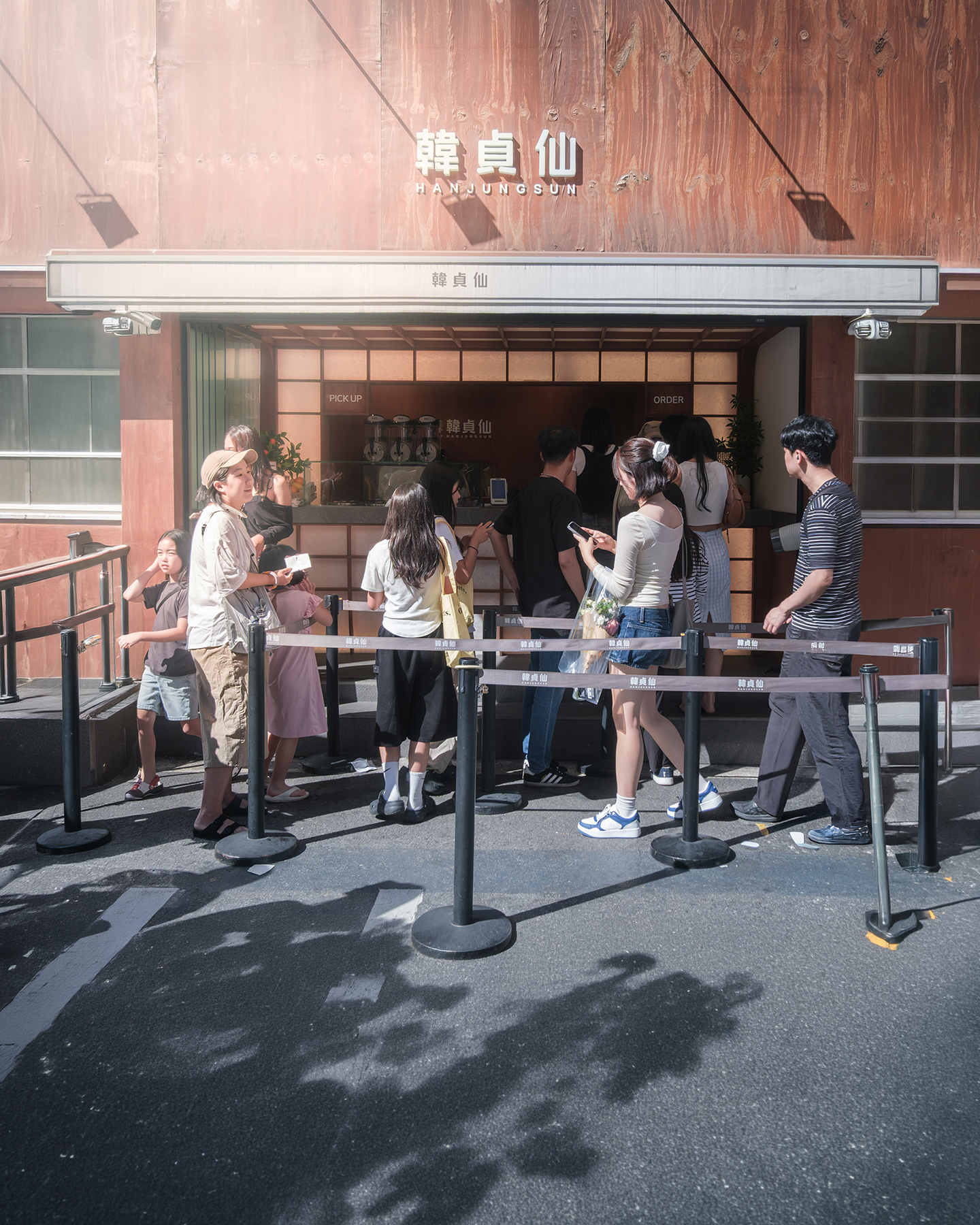 Shop - People queue outside a sunlit shopfront with wooden walls, metal barriers, and strong shadows across the street. - #Shop #Storefront #Street #Pedestrian #Passerby #StreetLife #StreetScene #StreetPhotography #UrbanLife #UrbanStreet