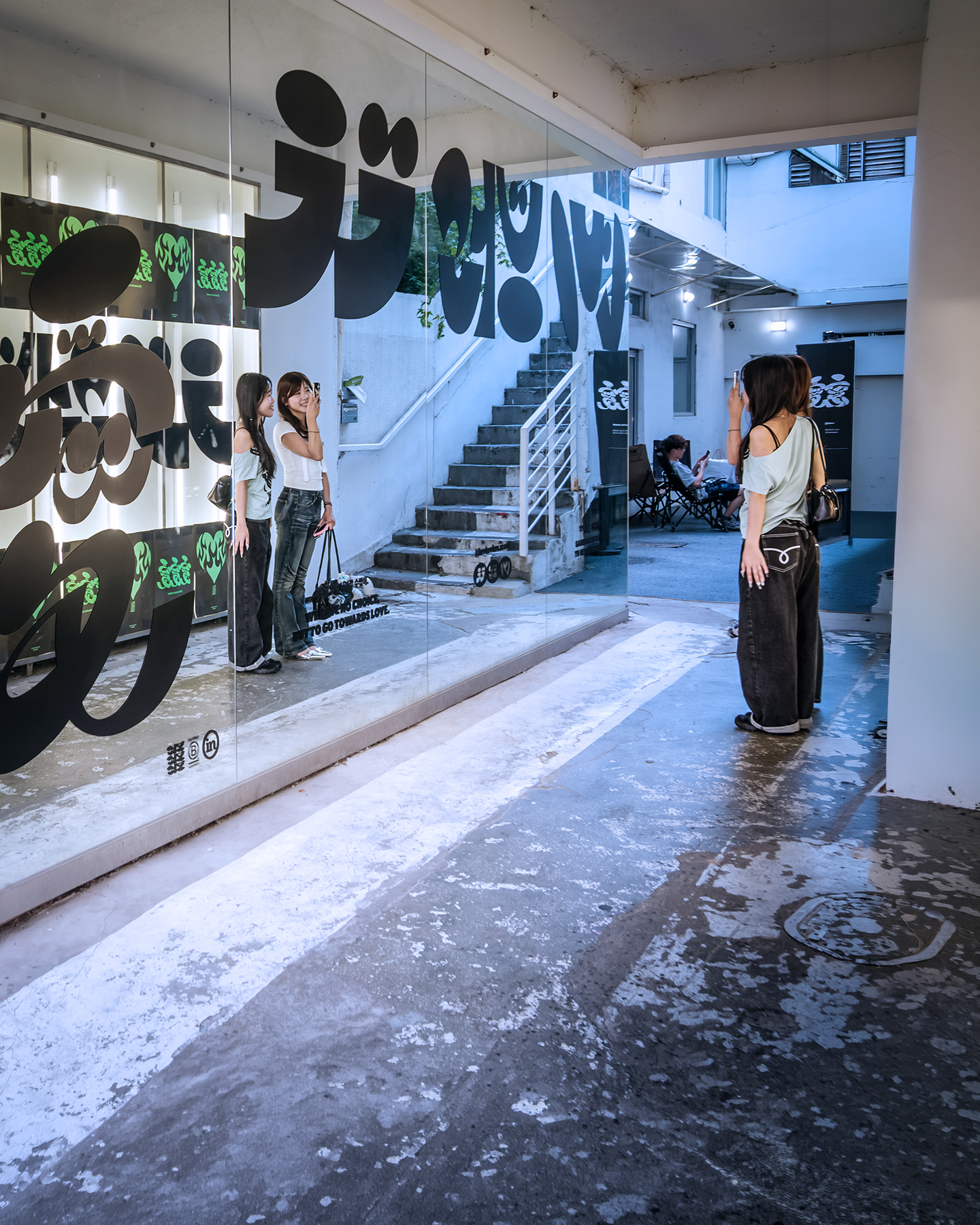 Mirror - Three women take photos beside a mirrored wall with bold black graphics in a cool-toned urban corridor. - #Mirror #StreetMirror #MirrorReflection #Reflection #Reflections #MirrorPhotography #ReflectionPhotography #Seongsu #SeongsuDong #SeongsuStreet