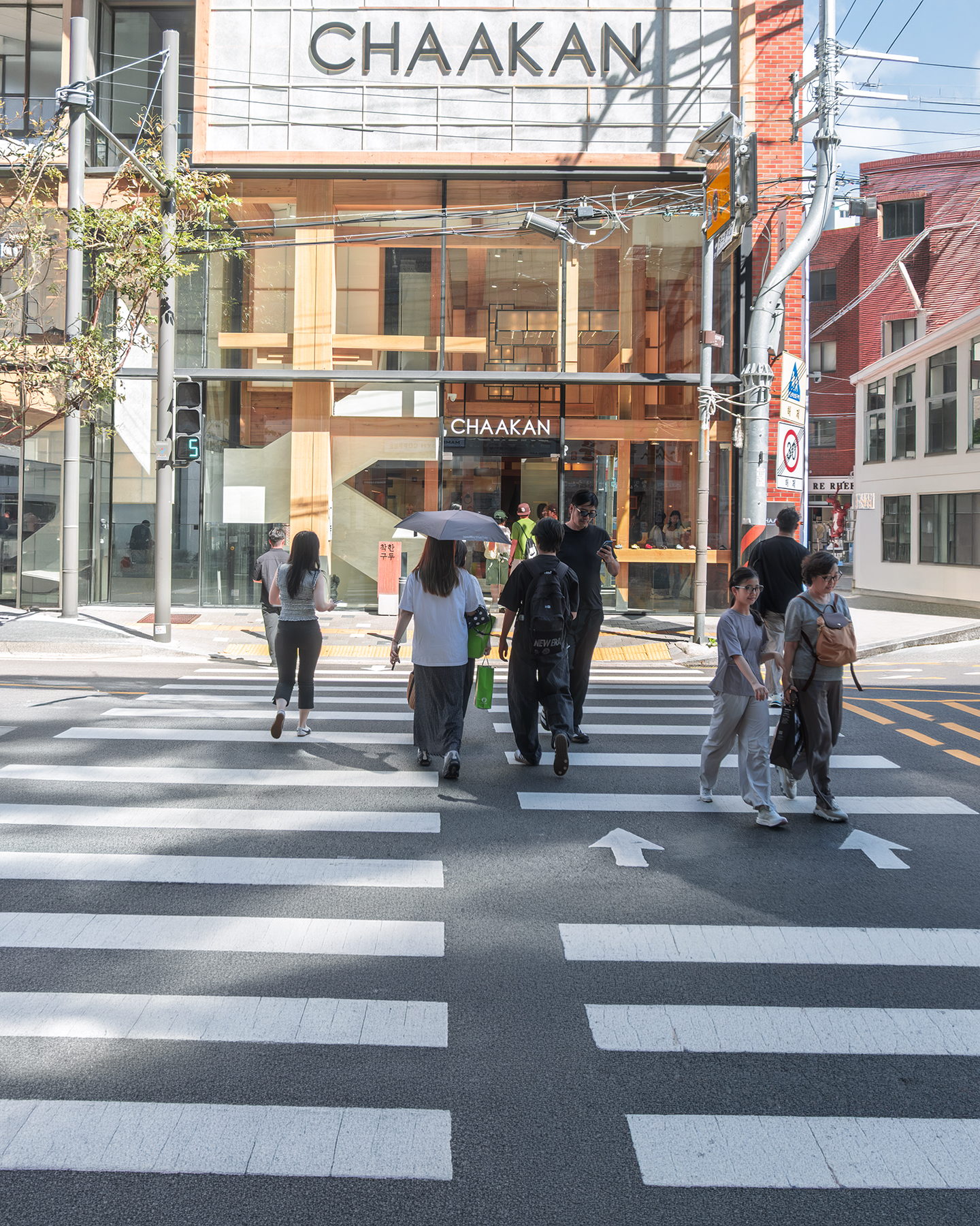 Crosswalk - Pedestrians cross a sunlit city crosswalk in front of a glass storefront, with bold white stripes and overhead wires framing the scene. - #Crosswalk #PedestrianCrossing #Street #Pedestrian #Passerby #StreetLife #StreetScene #StreetPhotography #UrbanLife #UrbanStreet