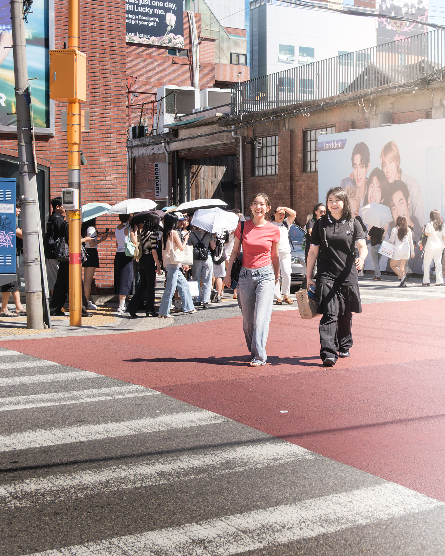 Crosswalk - Pedestrians cross a sunlit city street as umbrellas, brick facades, and bold crosswalk lines fill the busy urban scene. - #Crosswalk #PedestrianCrossing #Street #Pedestrian #Passerby #StreetLife #StreetScene #StreetPhotography #UrbanLife #UrbanStreet