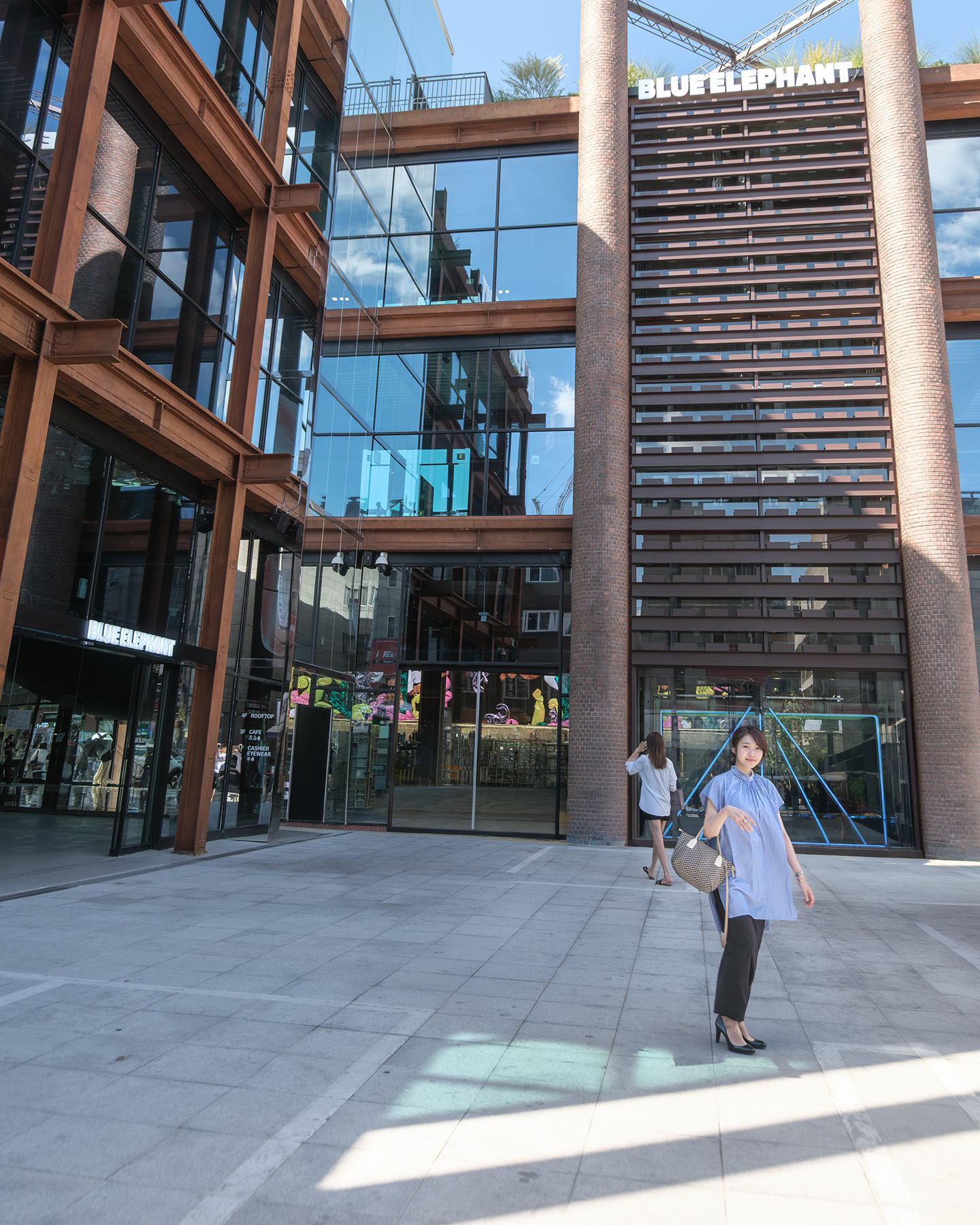 Blue Elephant - A woman stands in a sunlit plaza before a glass-and-brick building with Blue Elephant signage and strong architectural reflections. - #Seongsu #SeongsuDong #SeongsuStreet #Seoul #SouthKorea #Korea #SeoulStreet #SeoulStreets #StreetPhotography #SeoulStreetPhotography