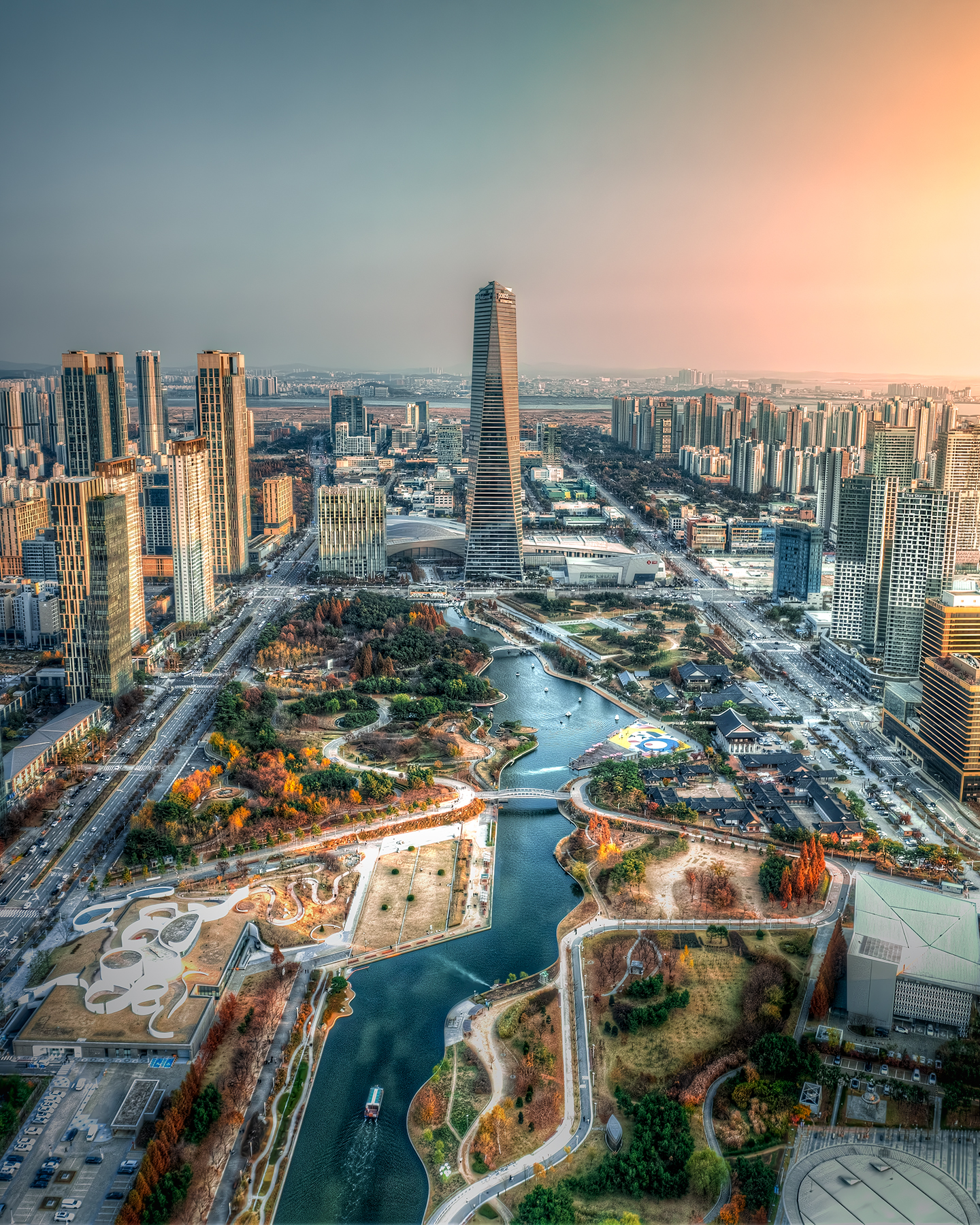 Songdo - A high-angle cityscape shows a winding canal, autumn parkland, and tall towers under warm hazy sunset light. - #Inchon #CentralPark #Songdo #SouthKorea