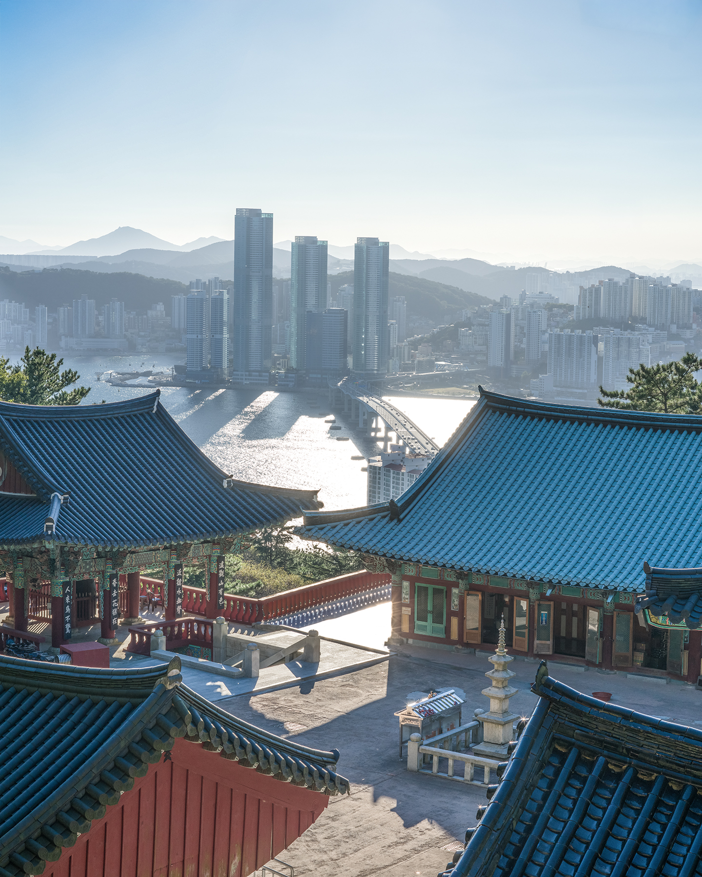 Bokcheon Temple - Blue-tiled temple roofs frame a hazy sunlit city, bridge, and waterfront with layered mountains in the distance. - #Busan #Sea #Building #SouthKorea