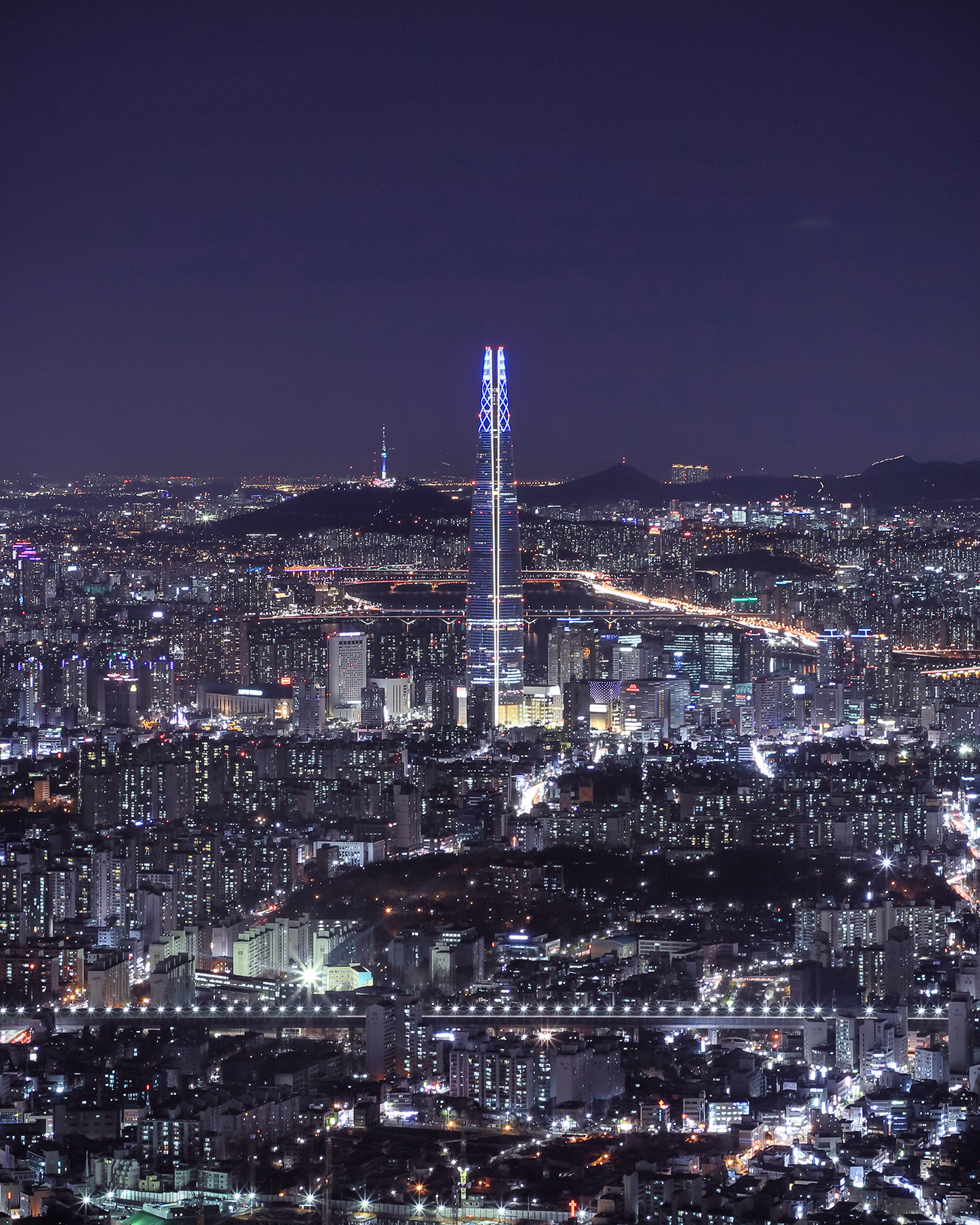Seoul - A brightly lit skyscraper rises above a dense nighttime cityscape, with bridges, roads, and distant hills under a dark sky. - #Seoul #Cityscape #NightPhotography #Skyline #UrbanLights #Skyscraper #LongExposure #Architecture #Nightscape #UrbanView
