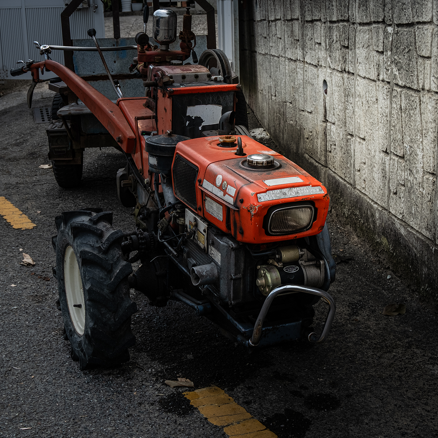 Cultivator - A weathered red cultivator stands in a narrow paved alley beside a textured concrete wall under subdued light. - #Cultivator #PowerTiller #WalkingTractor #TwoWheelTractor #FarmMachine #FarmMachinery #AgriculturalMachine #AgriculturalEquipment #FarmingEquipment #FarmEquipment