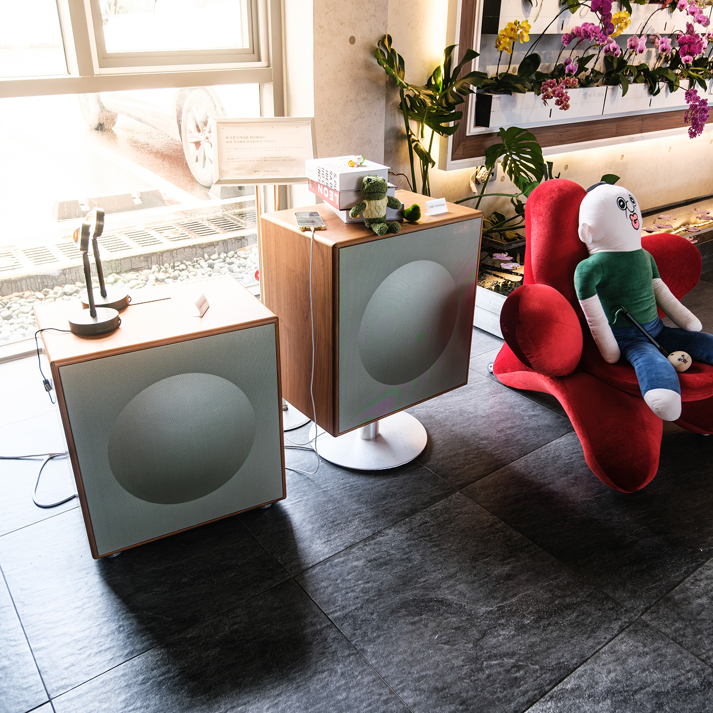 Hi Flowera - Sunlit flower shop interior with orchids, leafy plants, cube speakers, and a plush figure seated on a red chair. - #Cafe