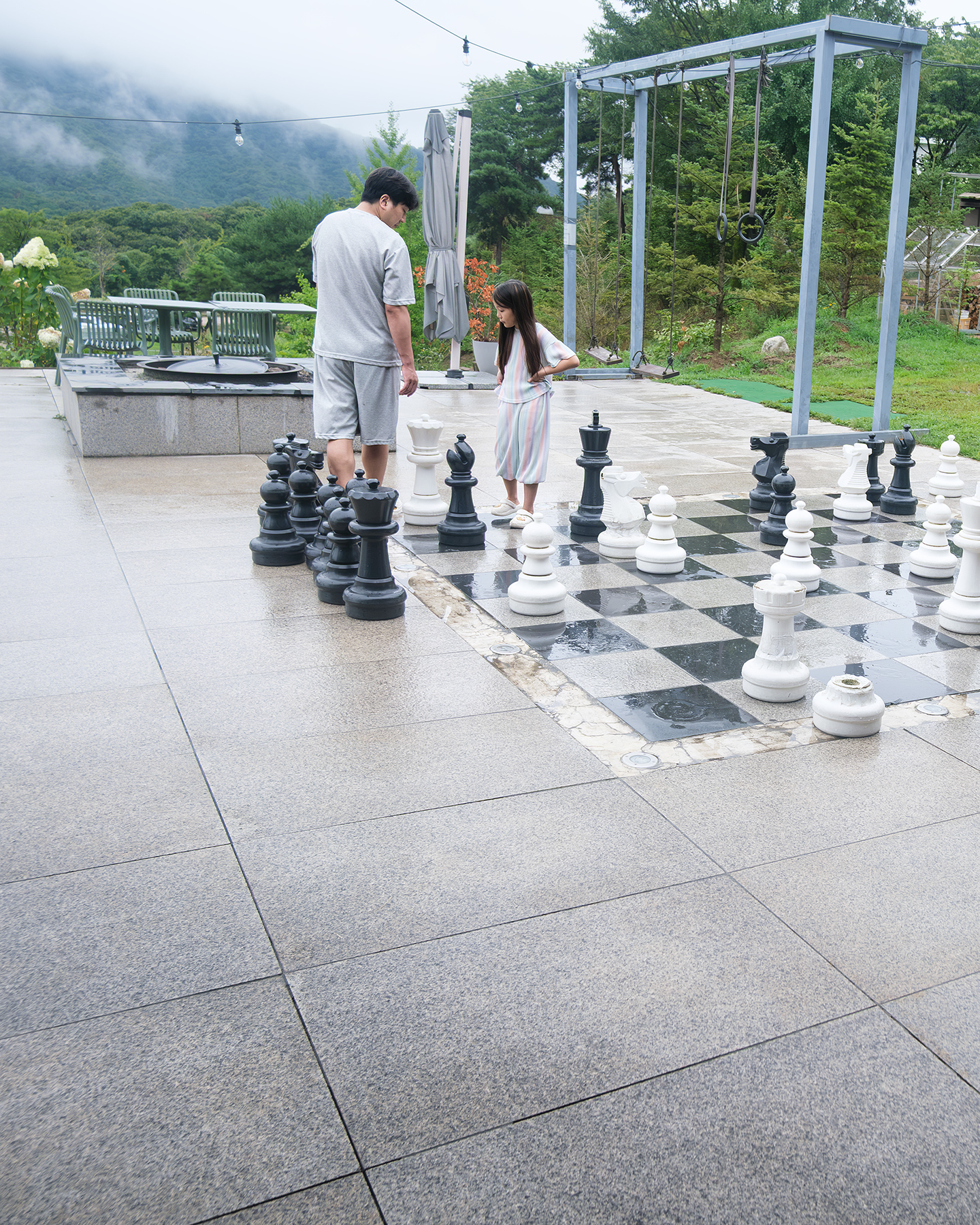 Birugae Botanical Garden - An adult and child stand beside oversized chess pieces on a wet garden terrace, with misty green hills in soft daylight. - #BirugaeBotanicalGarden #BirugaeCafe #BotanicalGardenCafe #GardenCafe #PlantCafe #GreenCafe #CafeInKorea #KoreanCafe #CafeTour #CafeHopping