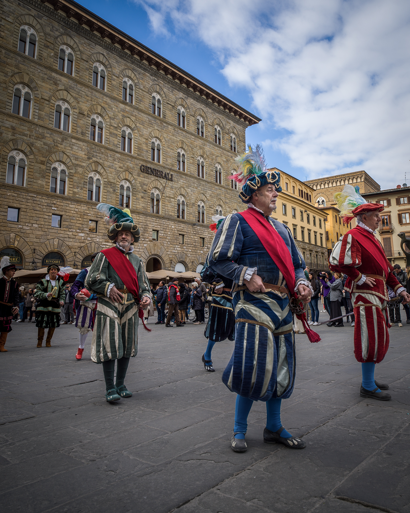 Florence - Costumed performers in feathered hats stand in a stone public square before ornate historic buildings under a partly cloudy sky. - #FlorenceFestival #FestivalInFlorence #FlorenceItaly #Firenze #FirenzeFestival #FlorenceEvents #ItalianFestival #ItalyFestival #FestivalVibes #StreetFestival