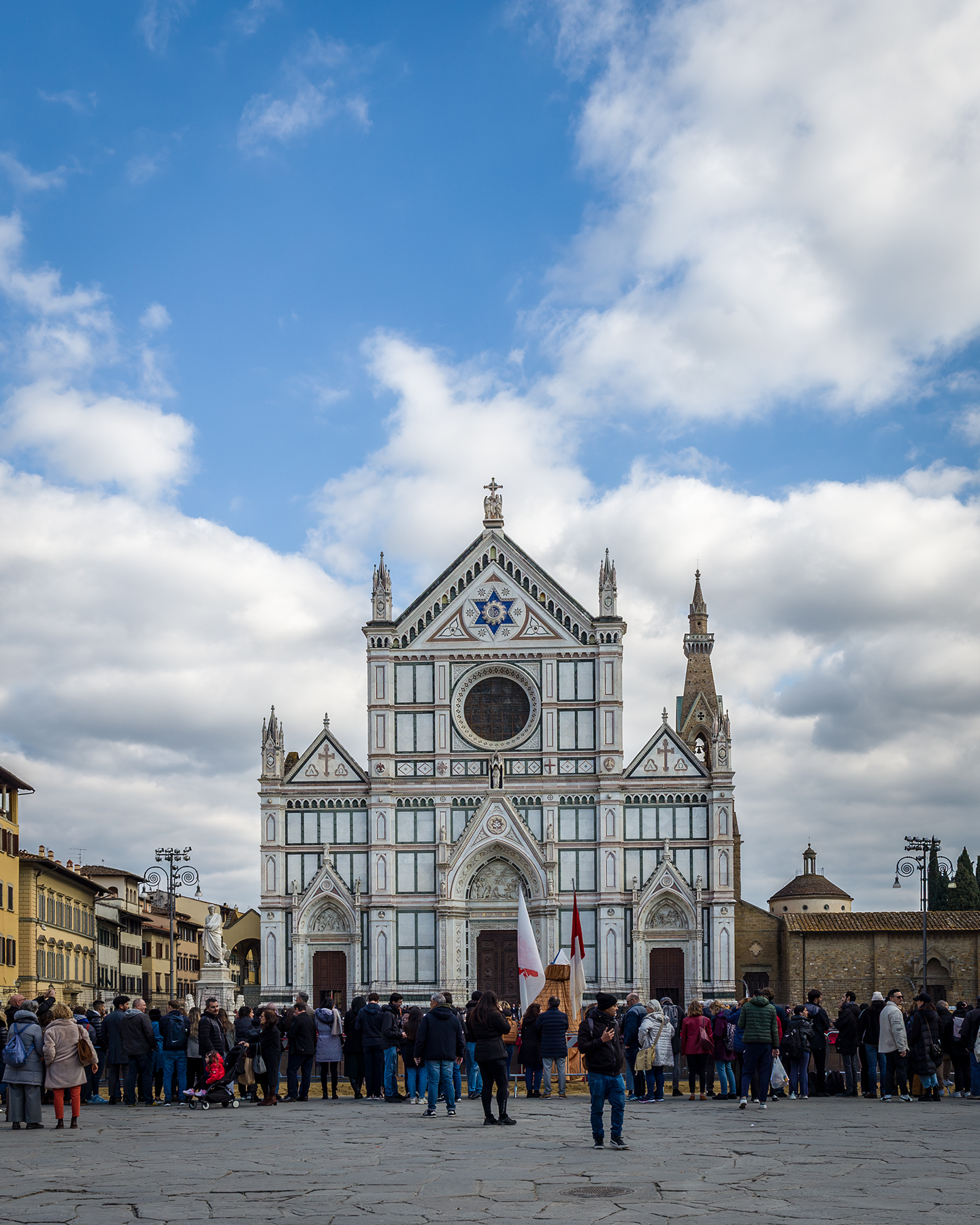 Santa Croce - A crowd gathers in a stone plaza before an ornate church facade under a bright blue sky with drifting clouds. - #SantaCroce #BasilicaDiSantaCroce #PiazzaSantaCroce #Florence #Firenze #FlorenceItaly #Tuscany #Toscana #Italy #VisitFlorence