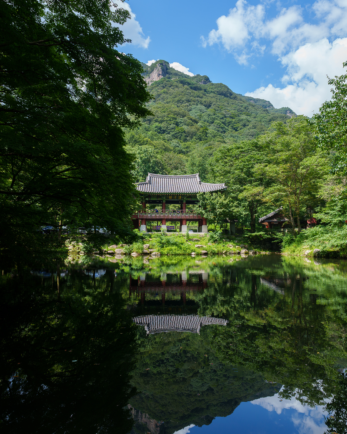 Baekyangsa - A traditional pavilion sits beside a still pond, reflected beneath lush forested mountains and a bright clouded sky. - #Baekyangsa #BaekyangsaTemple #BaekyangTemple #KoreanTemple #BuddhistTemple #KoreanBuddhism #TempleInKorea #KoreaTravel #TravelKorea #VisitKorea