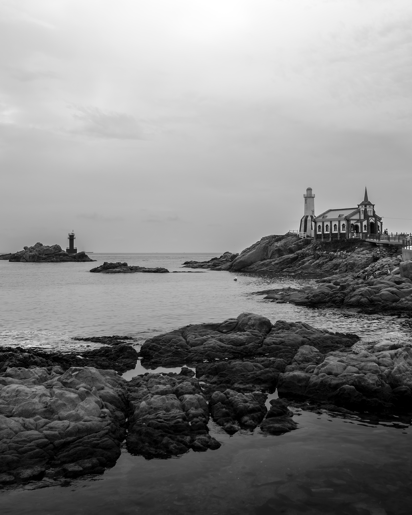 Dream Church - A black-and-white coastal scene shows a small church-like building on rocky cliffs, calm water, and a distant lighthouse under an overcast sky. - #BlackAndWhite #CoastalLandscape #RockyShore #Seascape #Chapel #Lighthouse #OvercastSky #CalmWater #MinimalComposition #FineArtPhotography