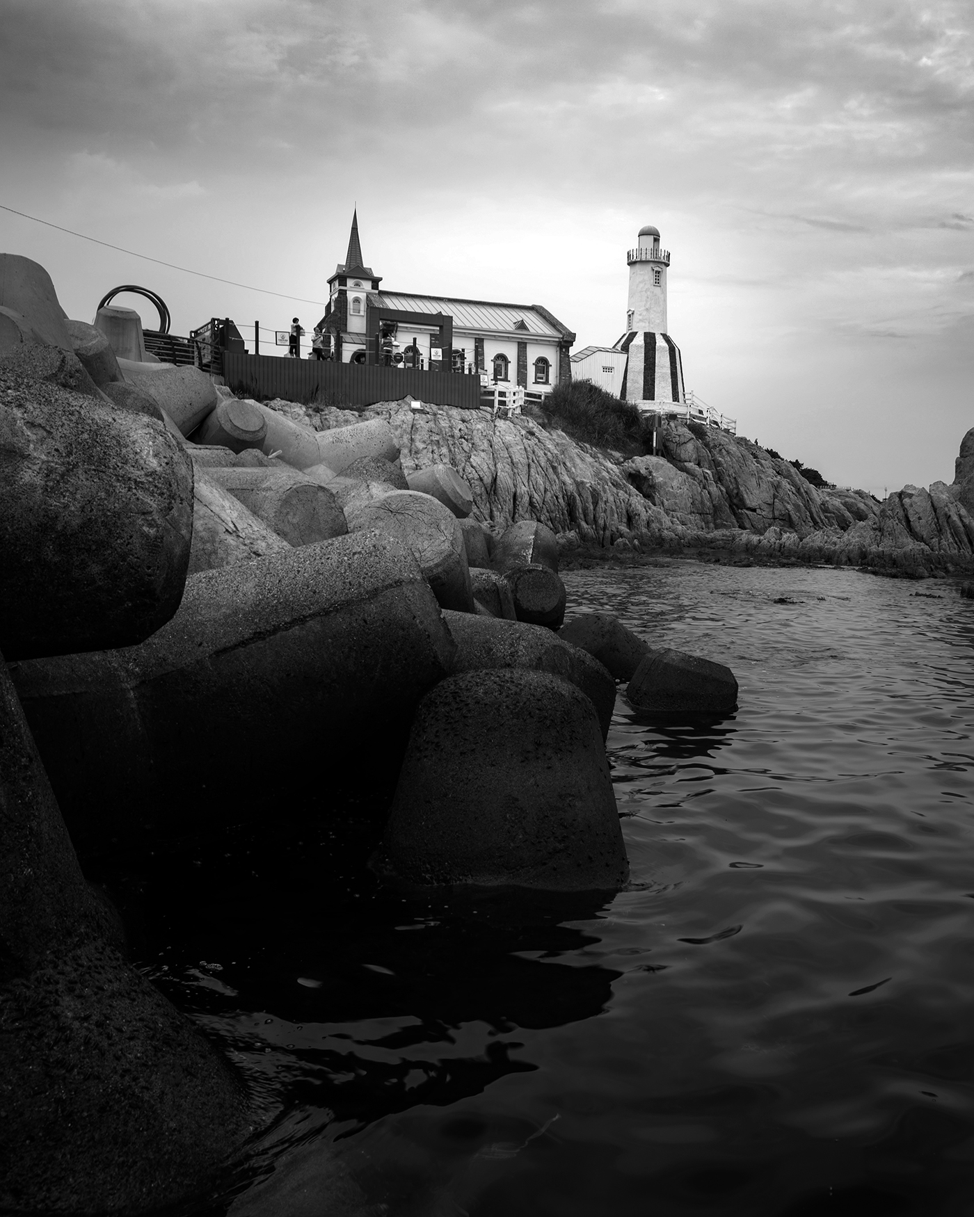 Dream Church - A monochrome coastal view shows a steepled building and lighthouse-like tower above rocks, tetrapods, and calm water under cloudy skies. - #BlackAndWhite #CoastalLandscape #Seascape #RockyShore #Steeple #LighthouseTower #CloudySky #Tetrapods #MonochromePhotography #Waterfront