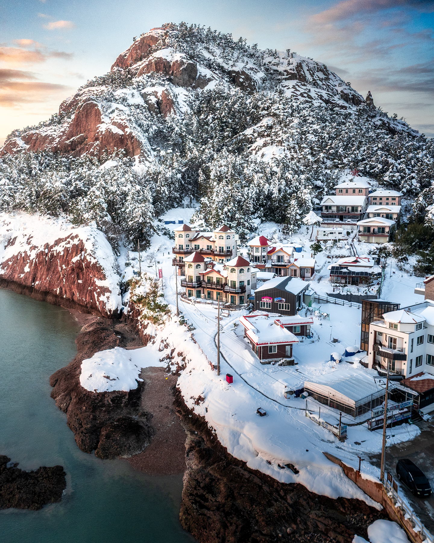 Daejangdo - Snow-covered houses sit below a rocky, forested coastal hill in warm low light beside calm blue-green water. - #Daejangdo #Daejangbong #Gunsan #GogunsanIslands #Jeollabukdo #SouthKorea #KoreaTravel #VisitKorea #WinterInKorea #WinterLandscape