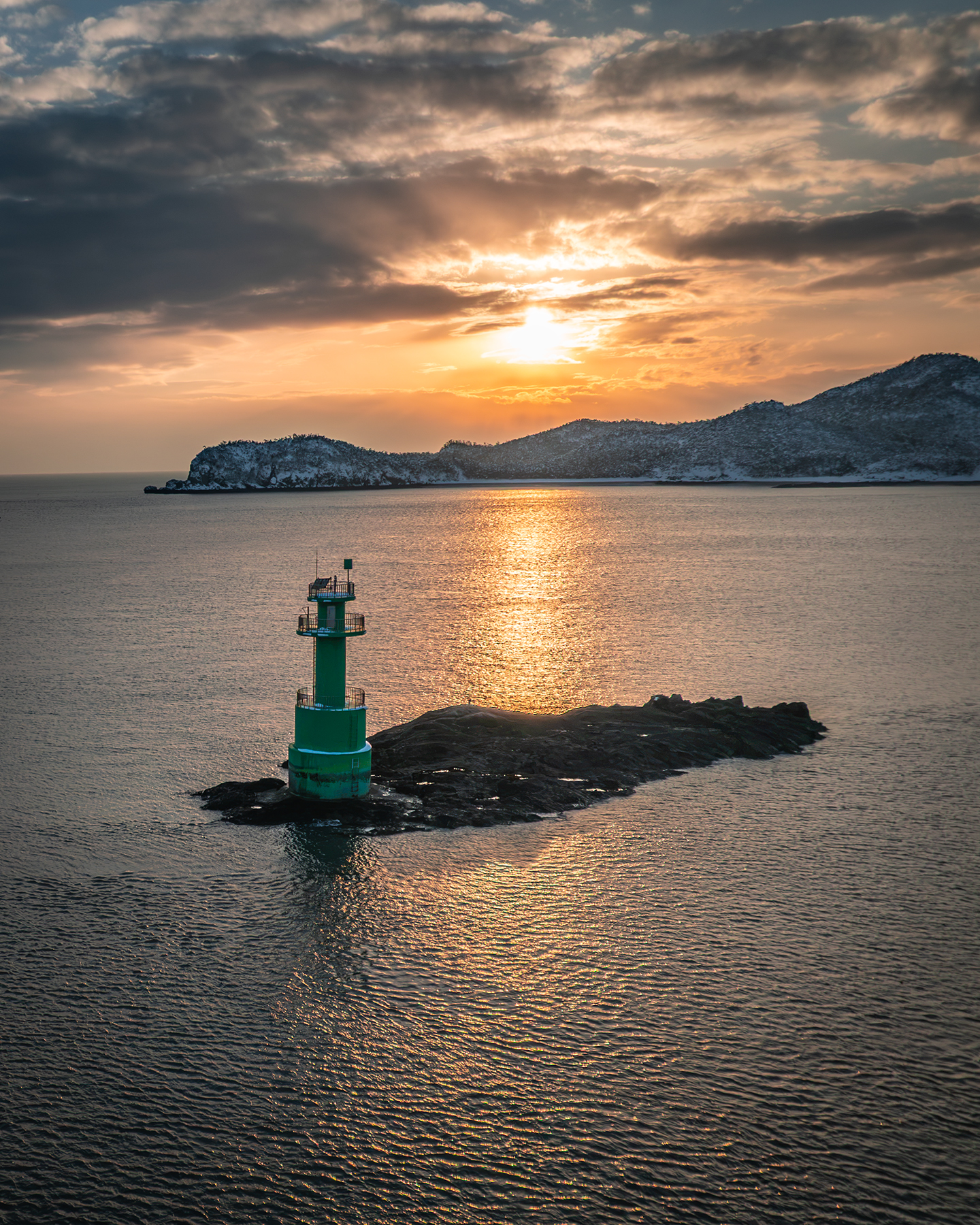 Daejangdo - A green lighthouse stands on a rocky islet as sunset light reflects across calm water beneath dramatic clouds and snowy hills. - #Daejangdo #Daejangbong #Gunsan #GogunsanIslands #Jeollabukdo #SouthKorea #KoreaTravel #VisitKorea #WinterInKorea #WinterLandscape