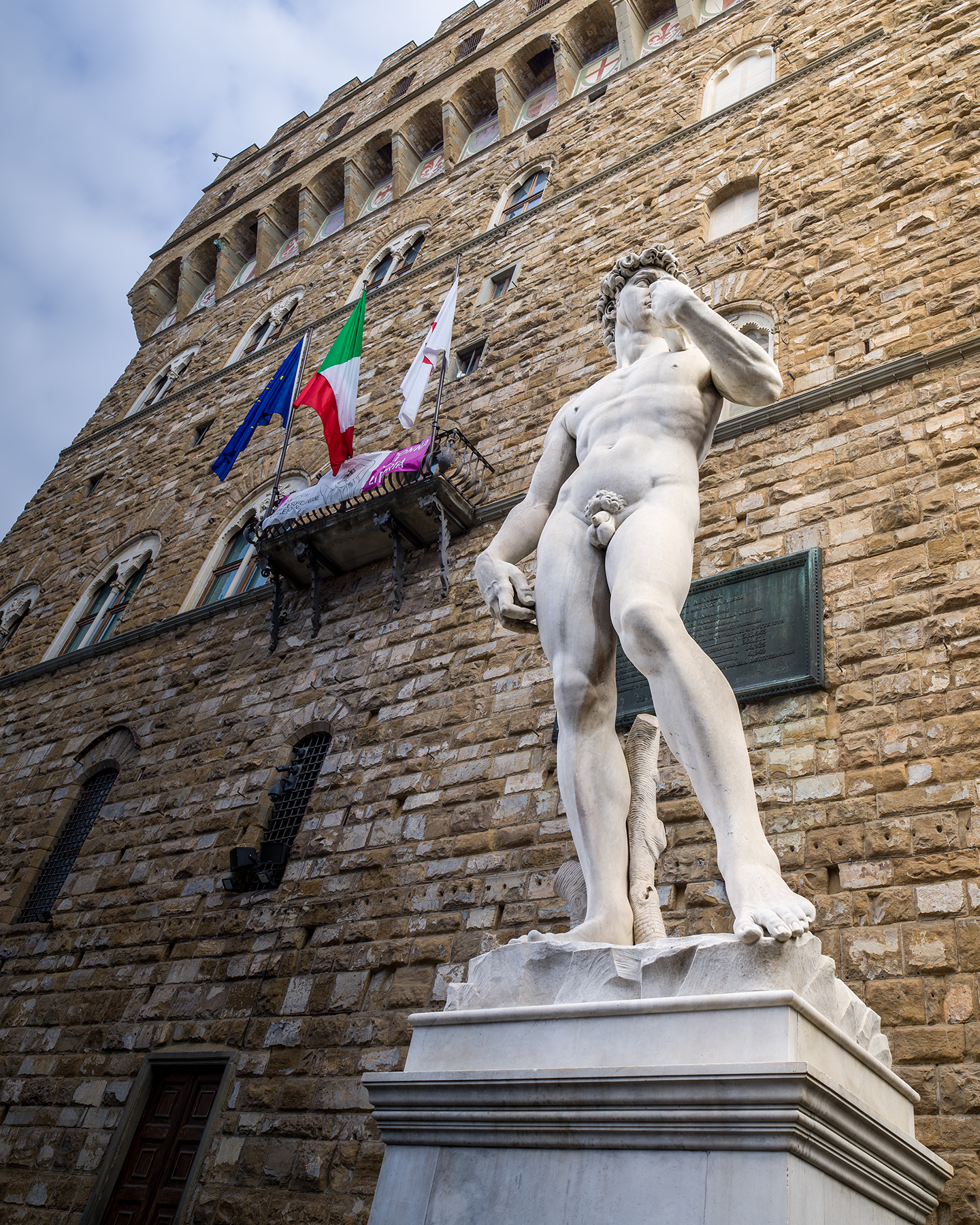 Florence - A marble nude statue stands before a tall rusticated stone facade with arched windows, flags, and soft cloudy daylight. - #David #DavidStatue #MichelangelosDavid #Michelangelo #MichelangeloBuonarroti #Florence #Firenze #FlorenceItaly #GalleriaDellAccademia #AccademiaGallery