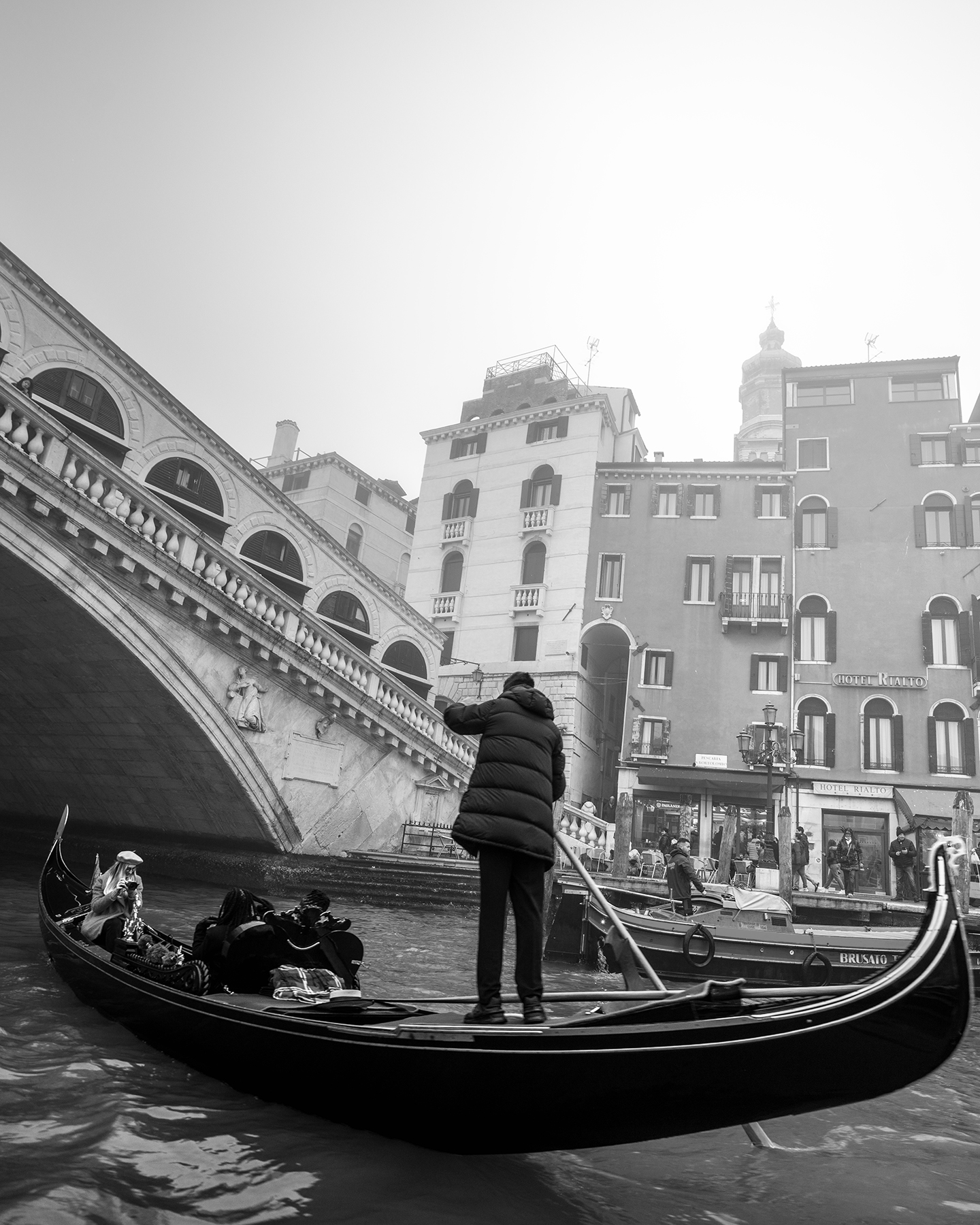 Venecia - A black-and-white gondola scene shows a gondolier on a canal beneath an arched bridge, with tall waterfront buildings in soft haze. - #BlackAndWhite #Gondola #Canal #Bridge #Architecture #StreetPhotography #TravelPhotography #UrbanScene #Waterfront #MoodyLight