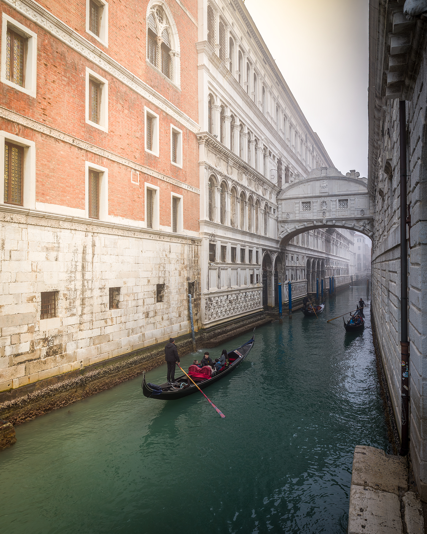 Venecia - Gondolas move along a narrow canal between ornate brick and stone buildings beneath a pale arched bridge in soft haze. - #Venice #Venezia #VeniceItaly #VeneziaItaly #Italy #VisitVenice #VisitVenezia #VisitItaly #ExploreVenice #ExploreVenezia