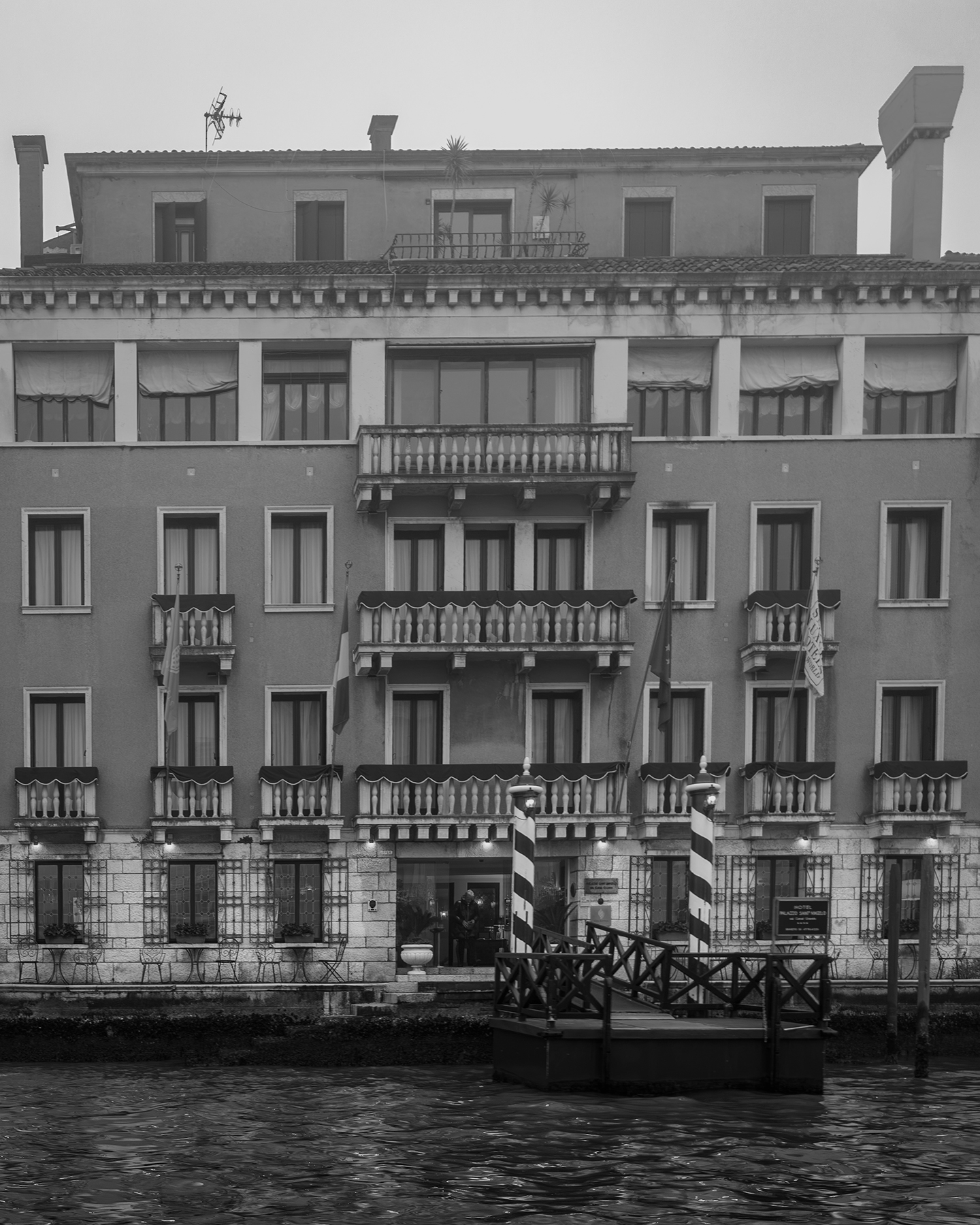 Venezia - A monochrome canal-side facade with rows of balconies, shuttered windows, striped mooring poles, and calm water under soft hazy light. - #Venezia #Italy