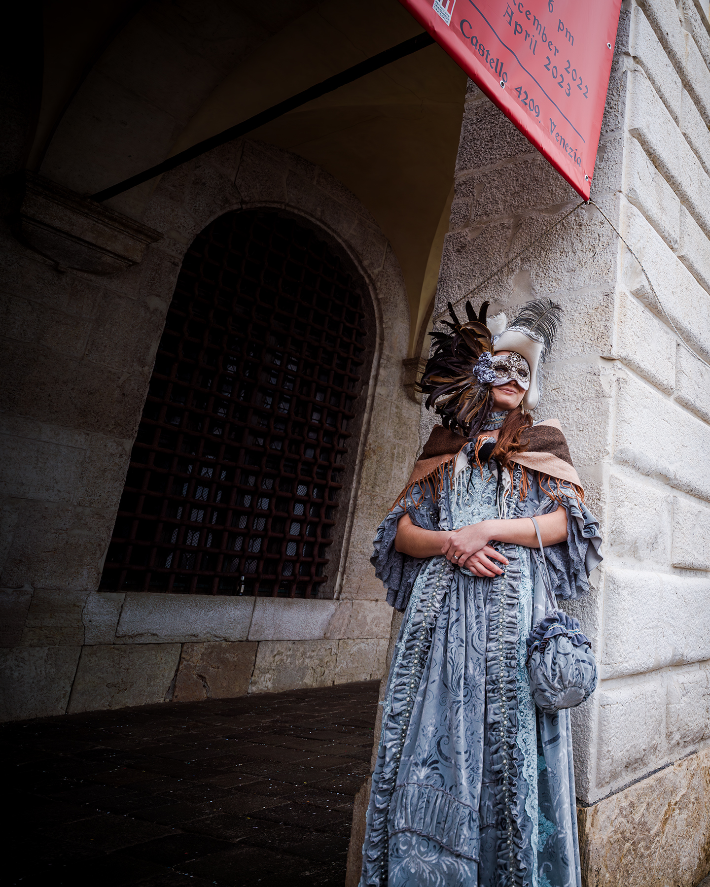Venezia Festival - A masked figure in an ornate blue costume stands by a stone arcade, lit from the side against a shadowed arched window. - #MaskedPortrait #OrnateCostume #StoneArcade #CarnivalMask #BlueDress #Feathers #ArchitecturalPortrait #SideLight #Shadows #HistoricArchitecture