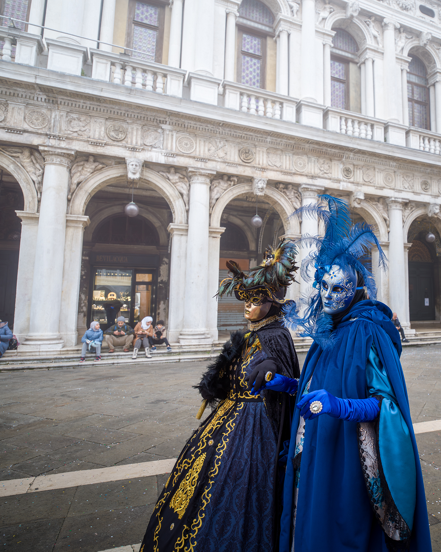 Venezia Festival - Two elaborately masked figures in blue and black costumes stand before ornate stone arches in soft daylight. - #MaskedFigures #CostumePhotography #OrnateArchitecture #StoneArches #BlueCostume #FeatheredMask #StreetPortrait #FestivalStyle #SoftLight #PublicSquare