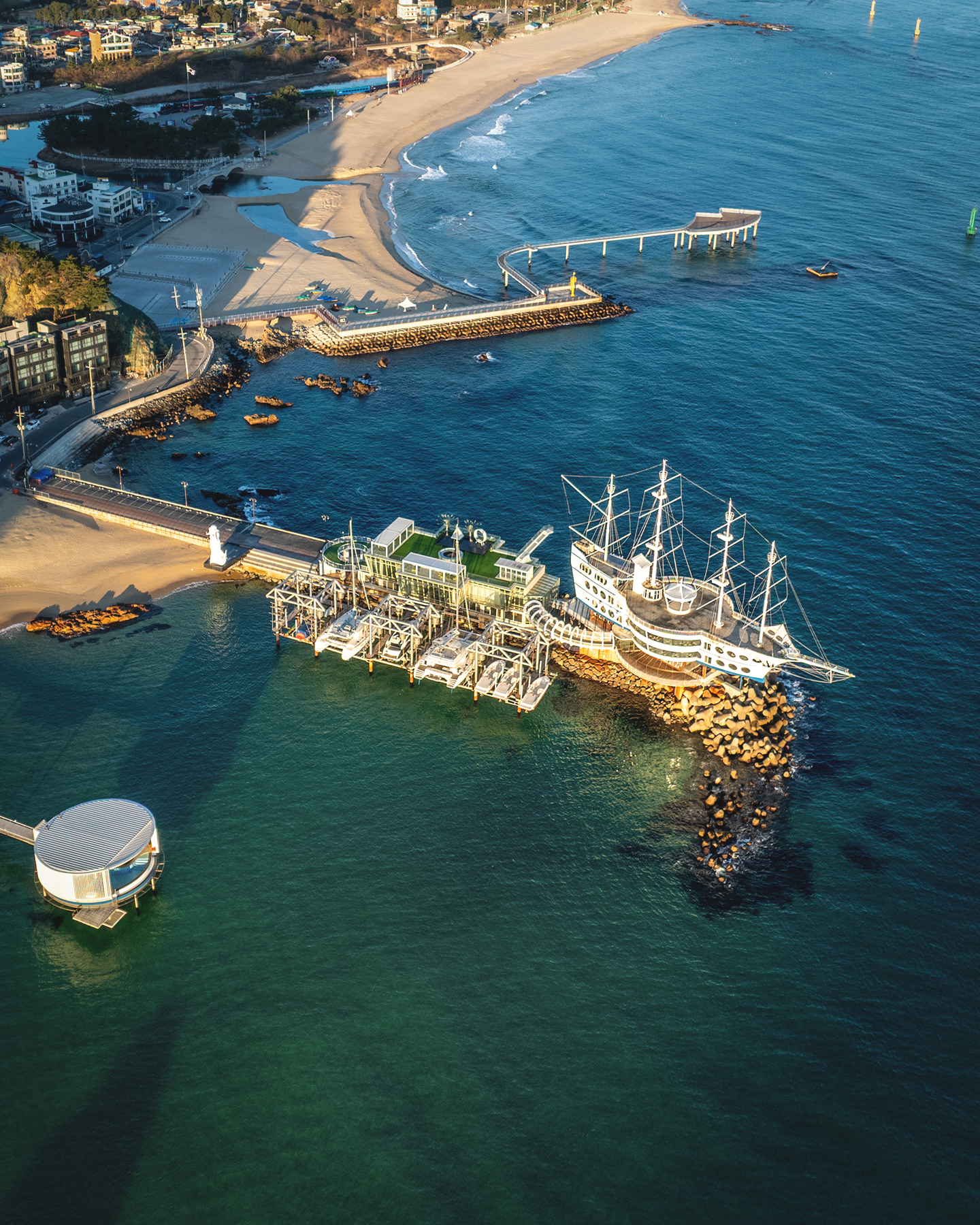 Jeongdongjin - An aerial view shows a sandy coast, blue sea, piers, and a ship-shaped waterfront building in warm low-angle light. - #Jeongdongjin #JeongdongjinBeach #Gangneung #GangneungTravel #GangneungTrip #Korea #SouthKorea #KoreaTravel #KoreaTrip #VisitKorea